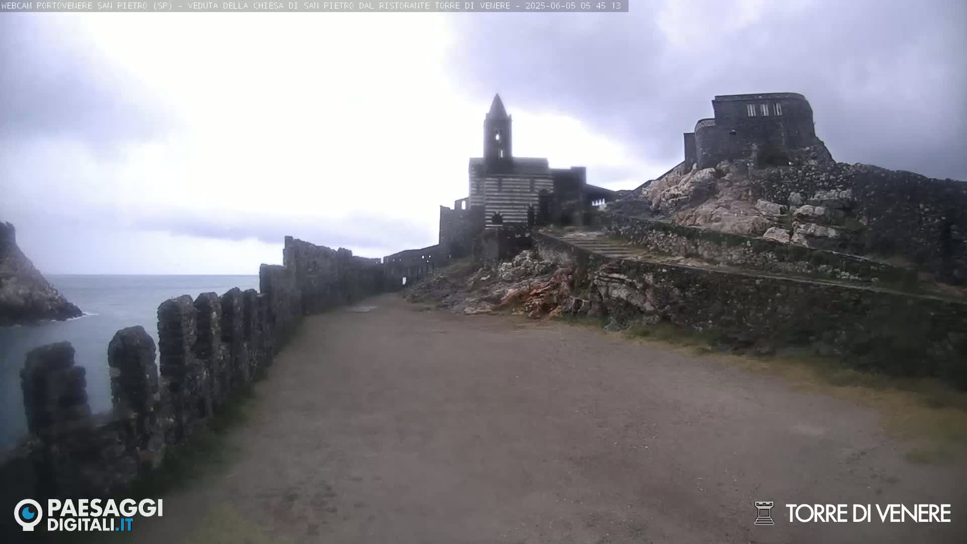 A stone path leads toward a striped church and stone buildings atop a rocky hill overlooking a calm sea under an overcast sky.