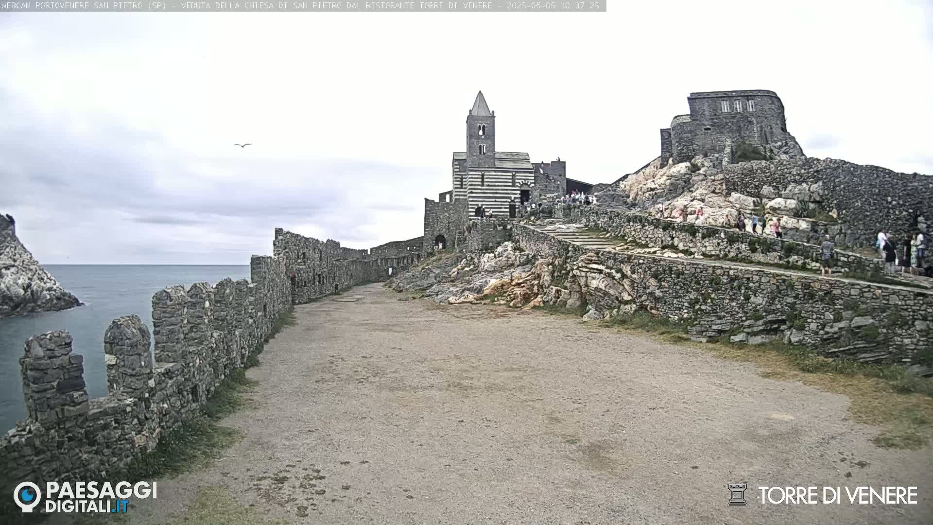 A stone pathway leads to a striped stone church and castle ruins perched on a rocky cliff overlooking a calm sea under an overcast sky.