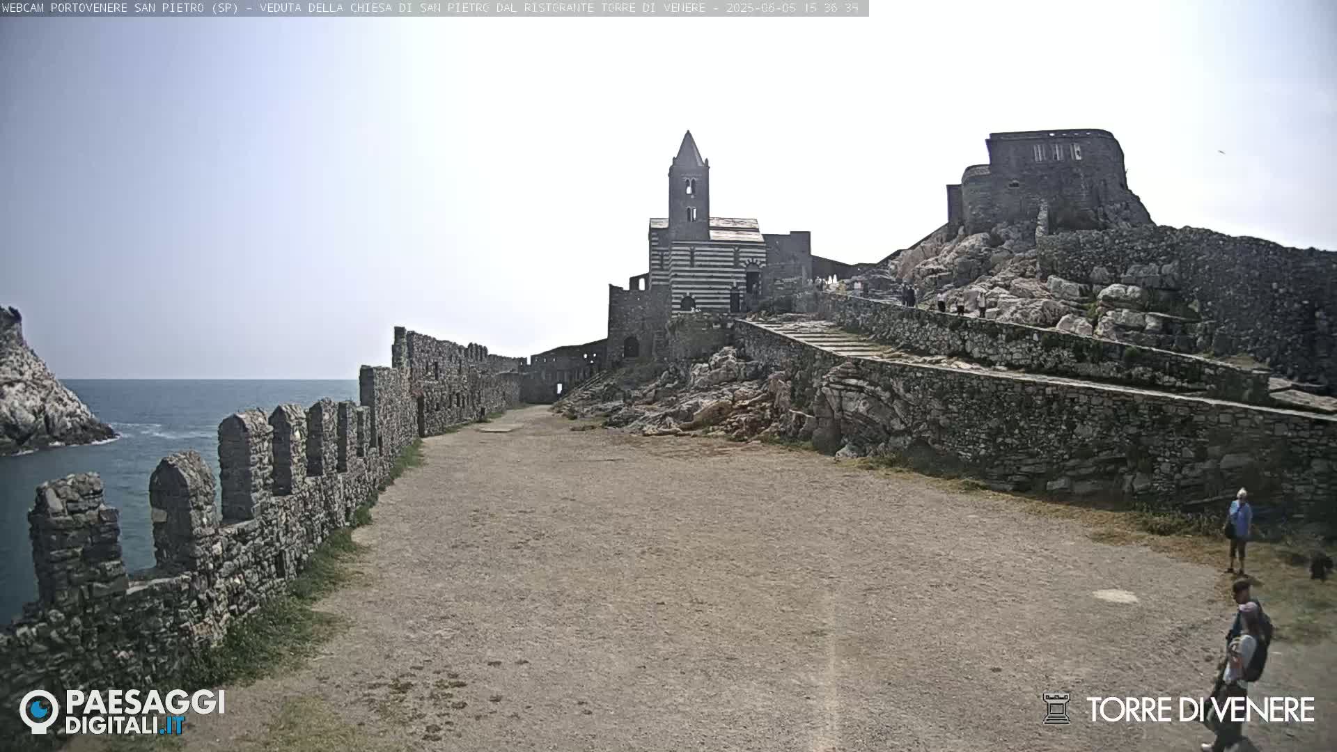 A stone church and castle ruins are situated on a rocky outcrop overlooking a calm sea under a bright, mostly clear sky.