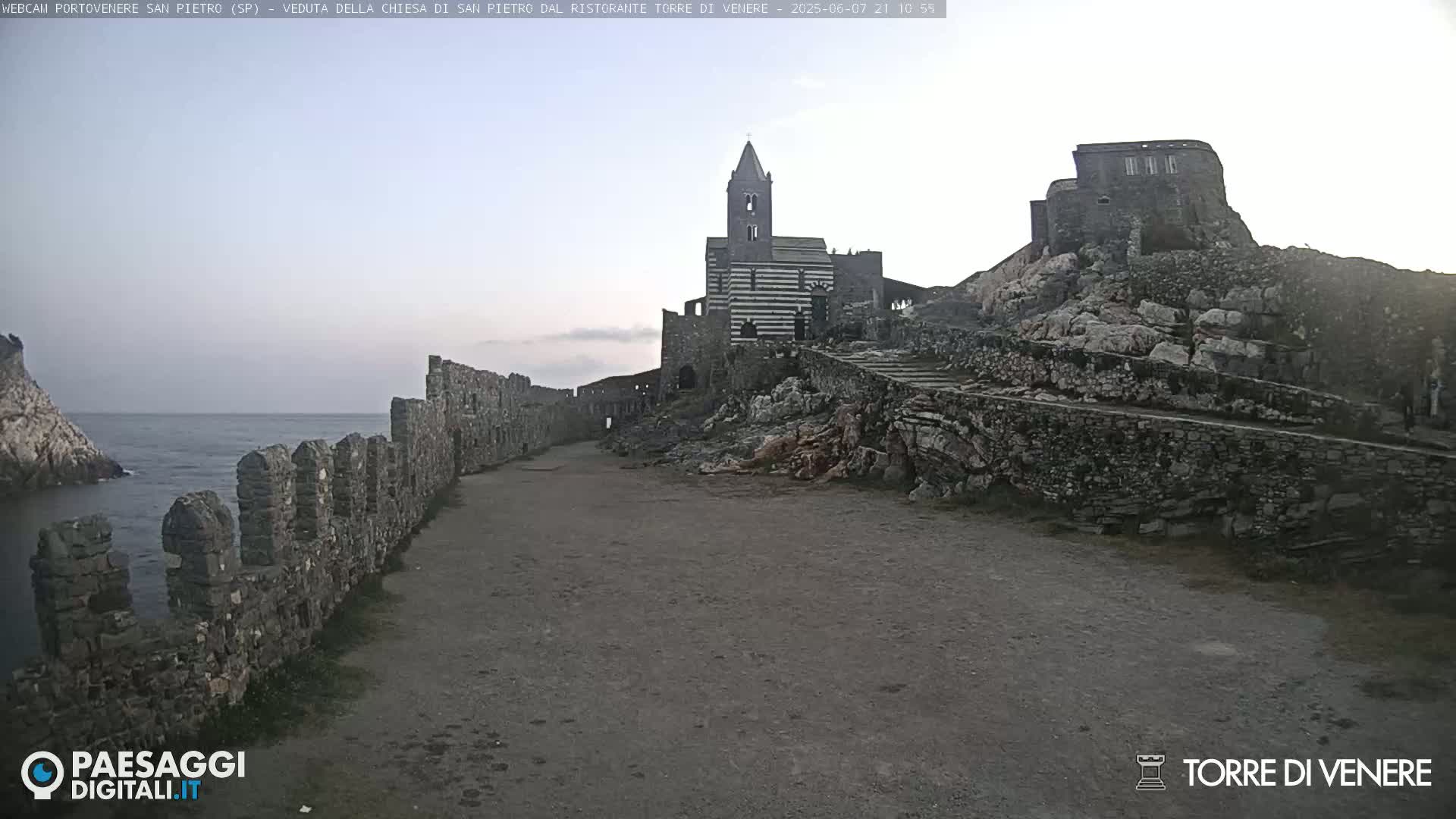 A stone pathway leads to a striped church and castle ruins overlooking a calm sea under a clear, twilight sky.
