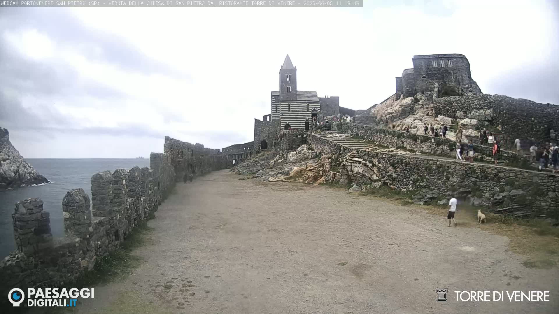 A rocky coastal area with ancient stone buildings, including a striped church and a fortress, is shown under an overcast sky.