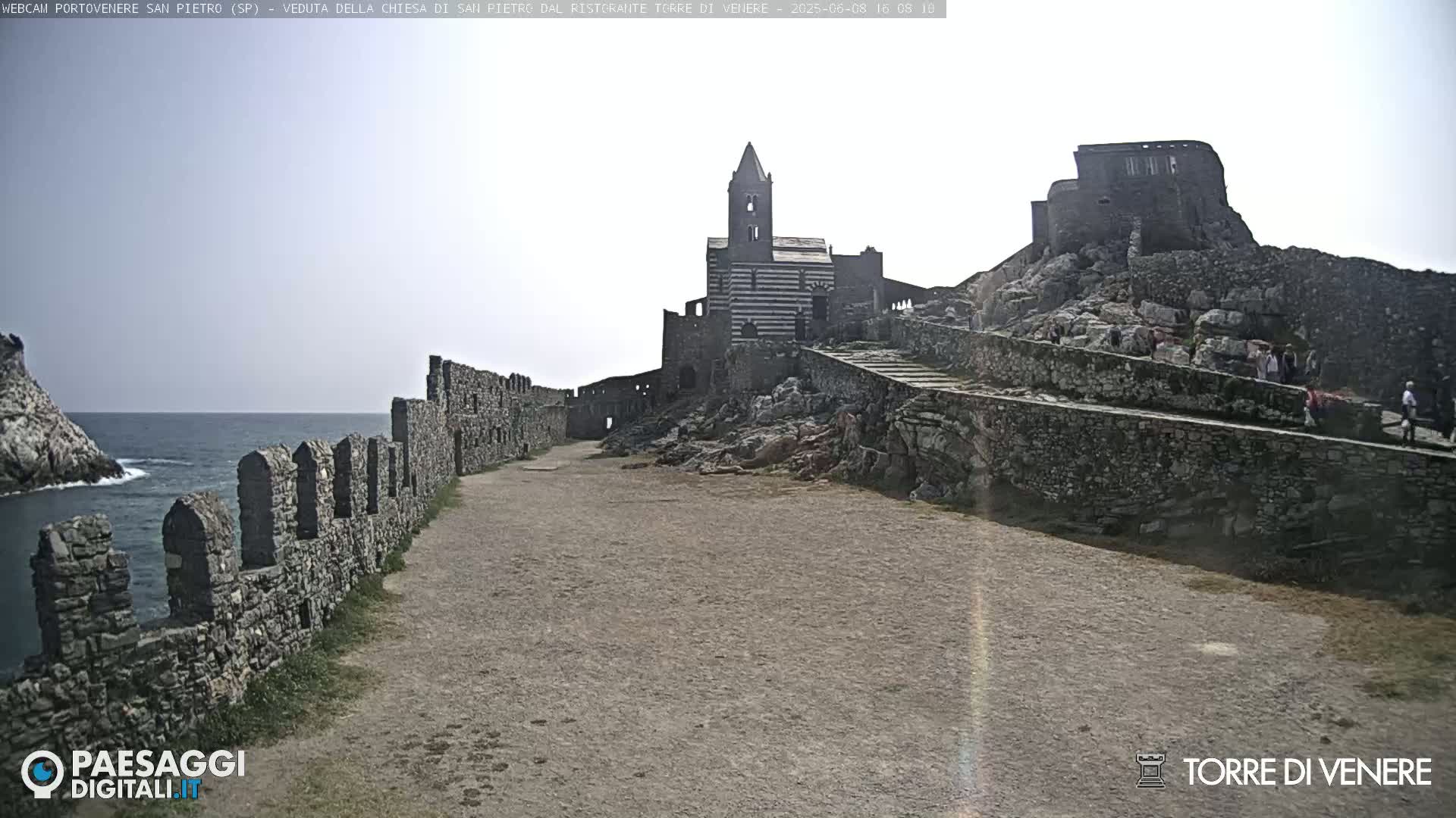 A stone pathway leads to a striped church and castle ruins overlooking a calm sea under a bright, mostly clear sky.