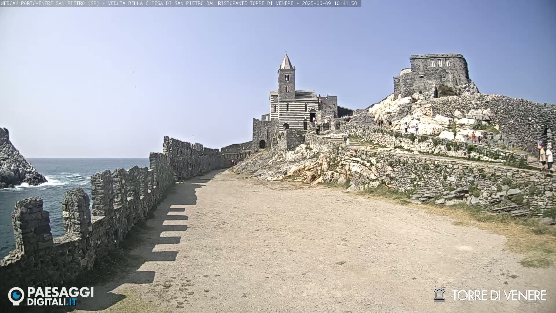 A stone pathway leads to a church and castle ruins overlooking a rocky coastline and ocean under a clear sky.