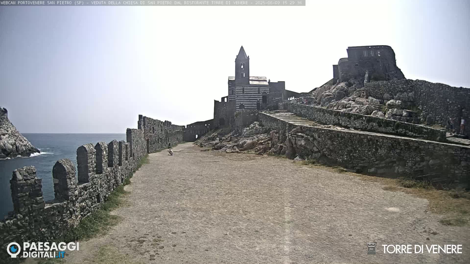 A stone pathway leads toward a striped church and castle ruins overlooking a calm ocean under a bright, mostly clear sky.