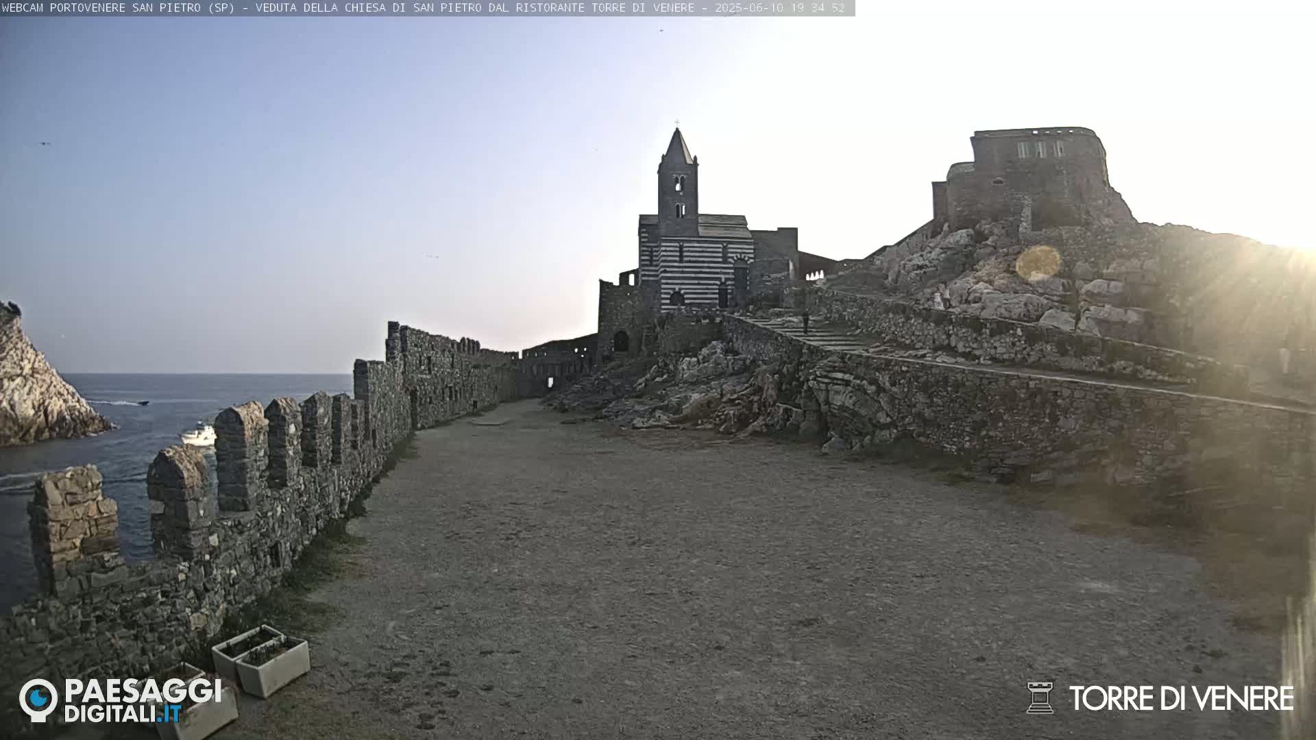 A stone church and castle ruins stand on a rocky outcrop overlooking a calm sea under a mostly clear sky.