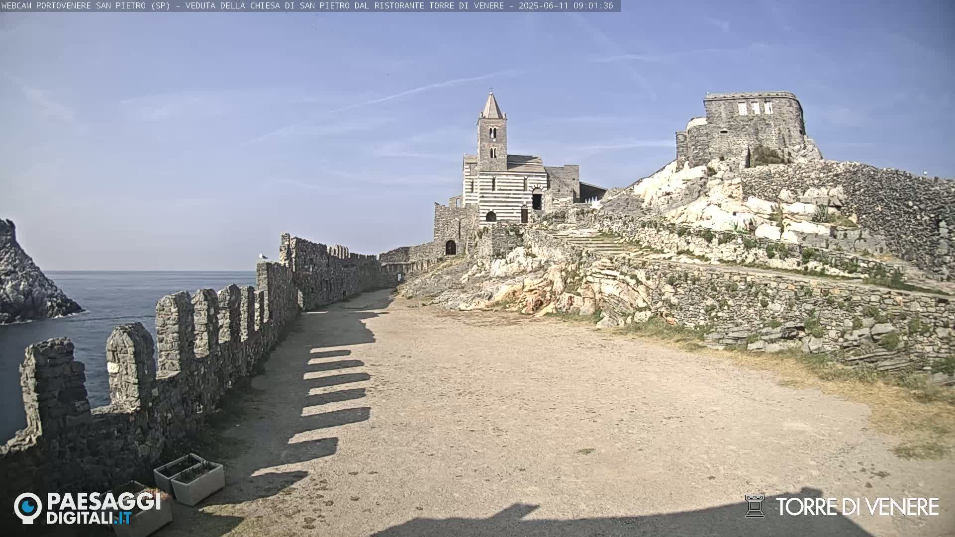 A stone pathway, bordered by a low wall, leads to a striped stone church situated atop a rocky hill overlooking a calm ocean.