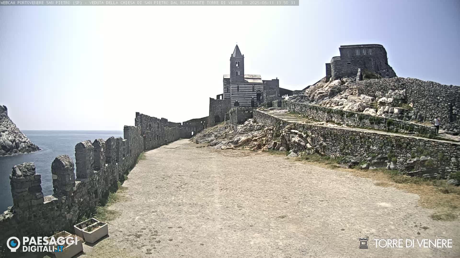 A stone pathway leads to a striped church and castle ruins perched on a rocky cliff overlooking a calm ocean under a mostly clear sky.
