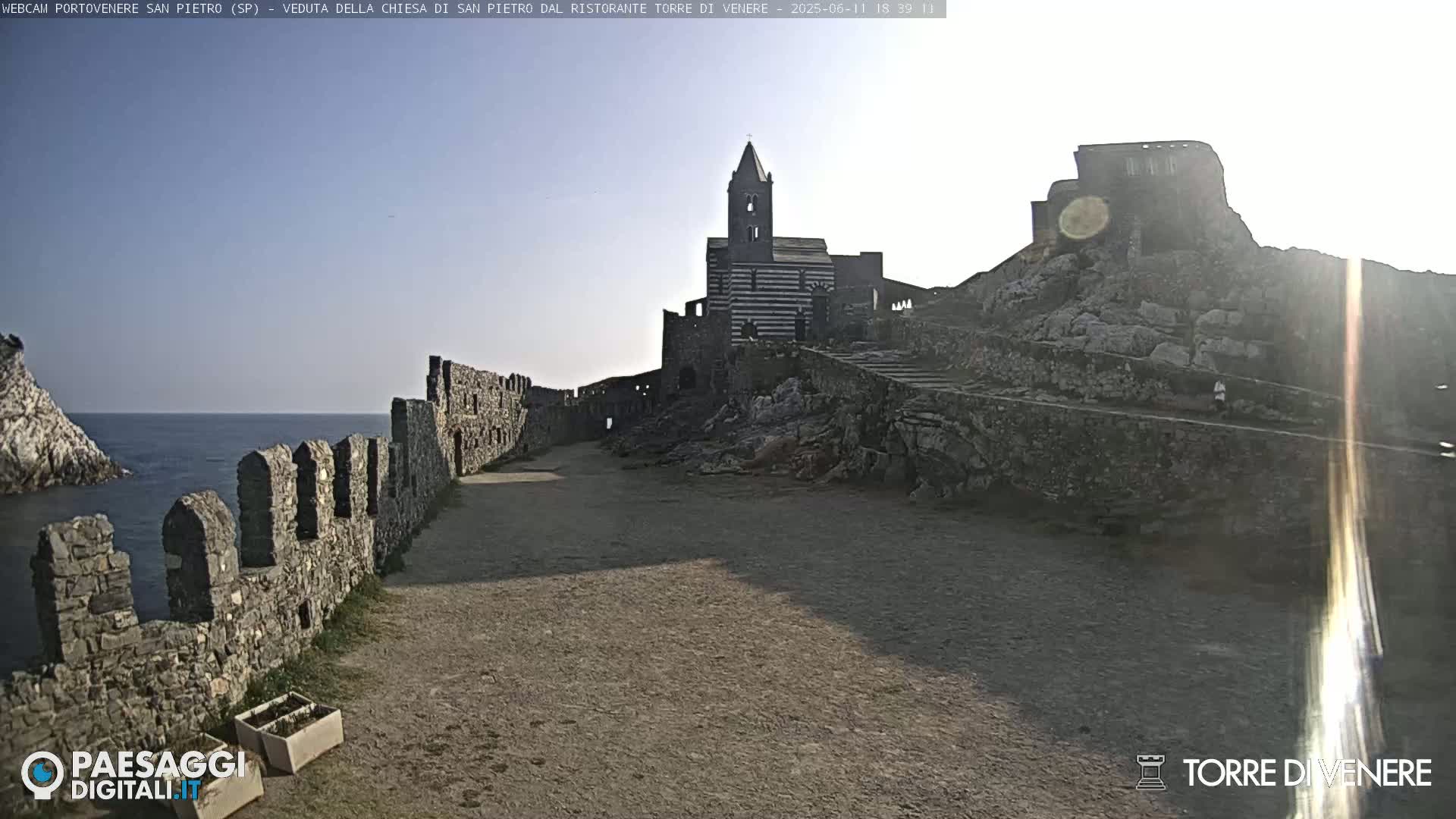 A stone pathway leads to a striped church situated on a rocky hill overlooking the sea under a mostly sunny sky.
