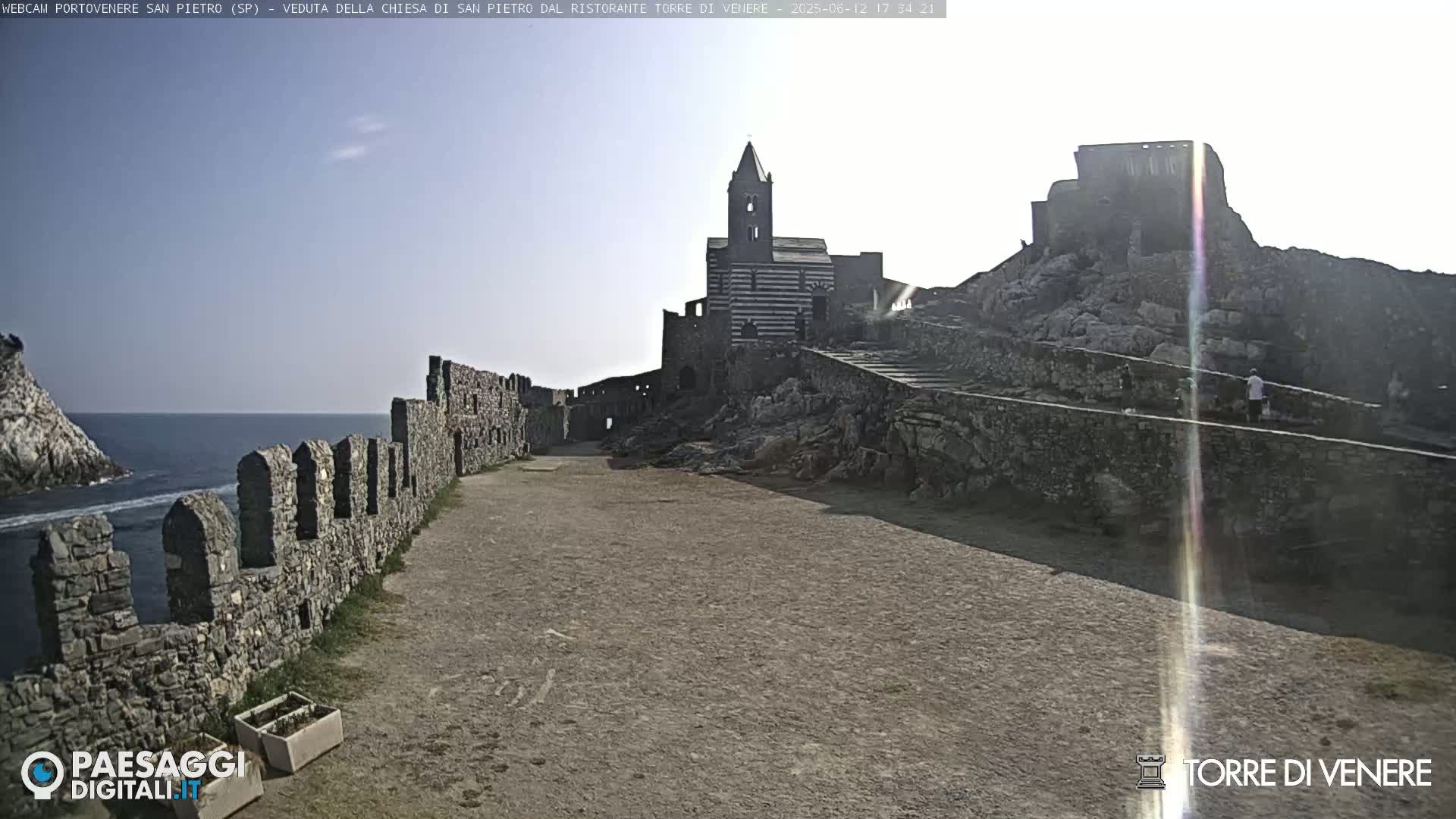A stone pathway leads toward a striped church and castle ruins overlooking a calm sea under a mostly clear sky.