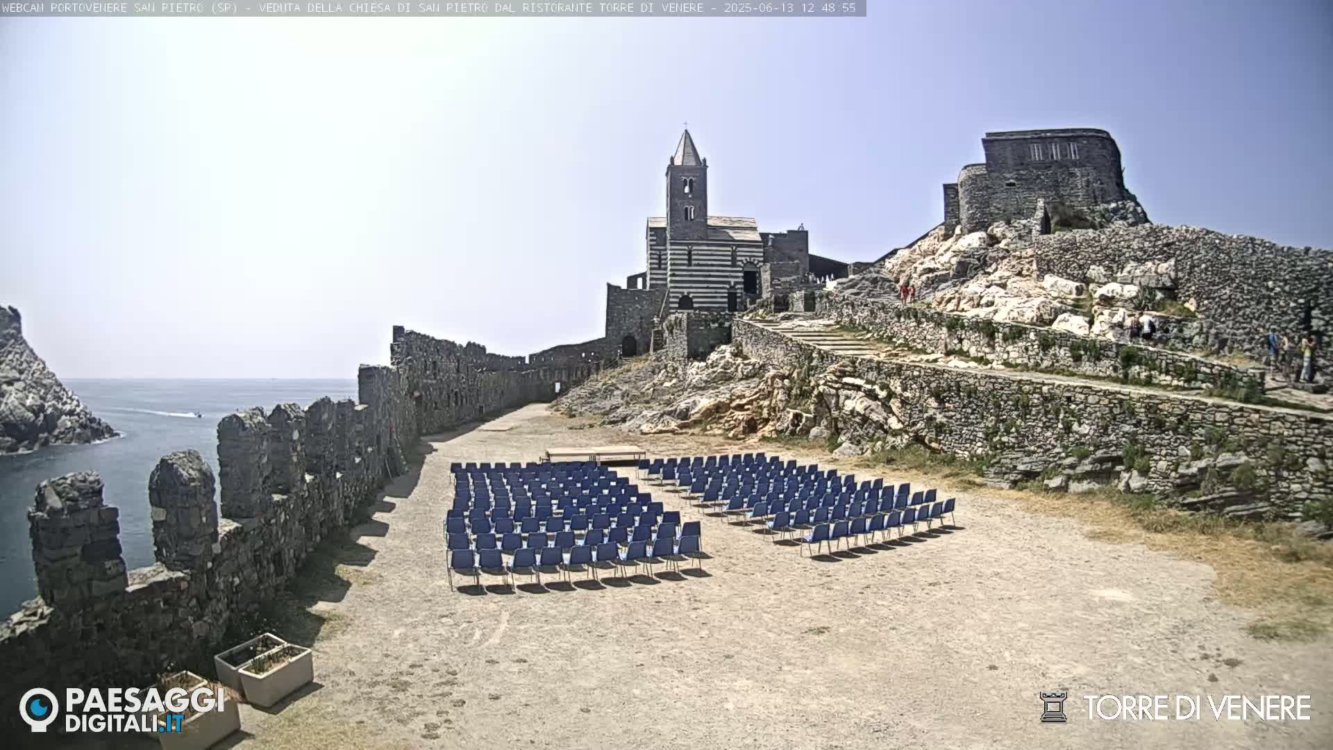A stone church and castle ruins overlook an empty outdoor seating area on a sunny day, with a calm sea visible in the distance.