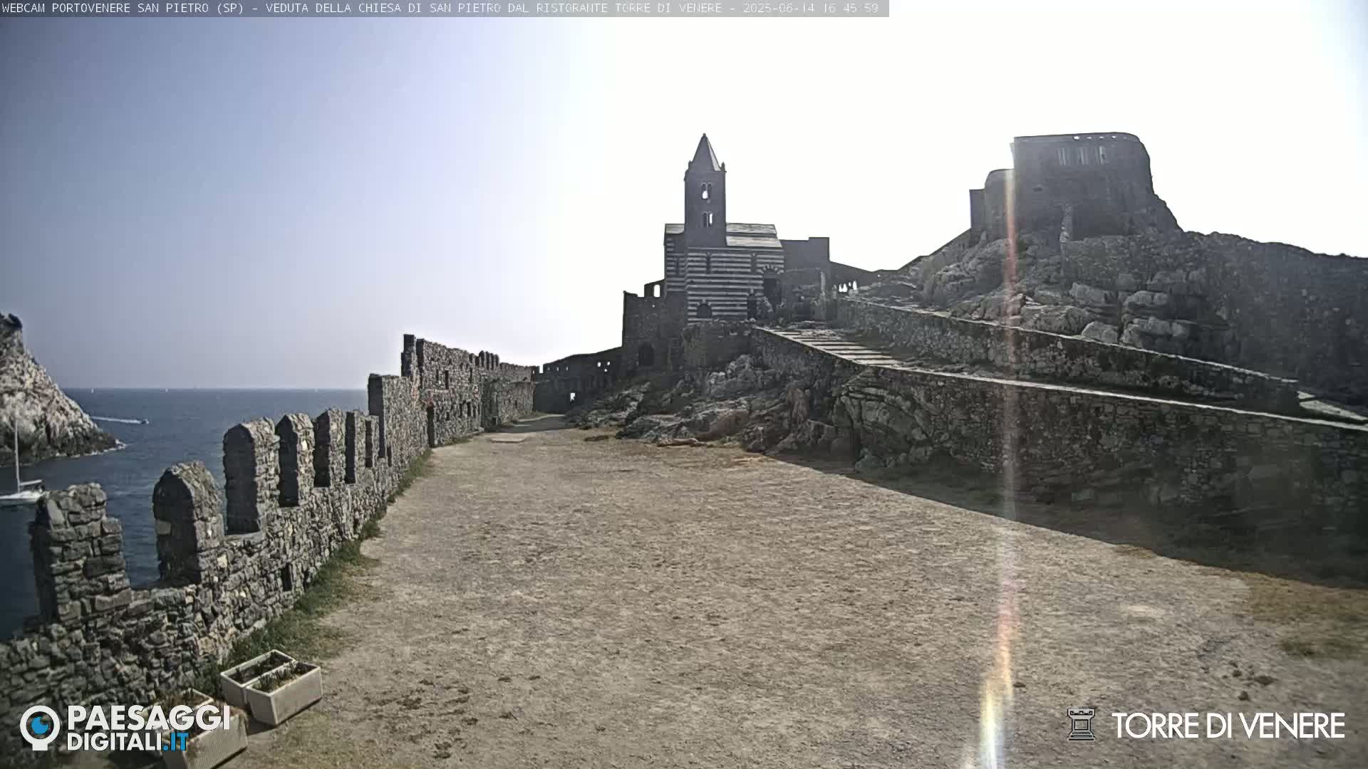 A stone pathway leads to a striped church and castle ruins overlooking a calm sea under a bright, sunny sky.