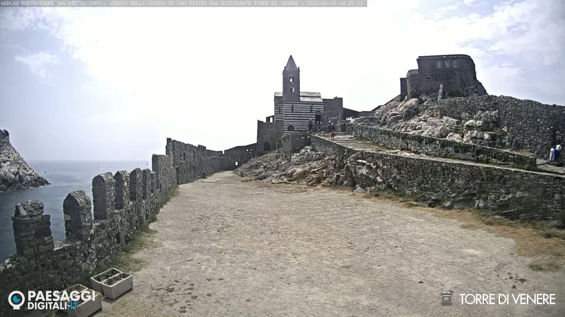 A stone pathway leads to a striped stone church and stone structures atop a rocky cliff overlooking a calm ocean under a partly cloudy sky.