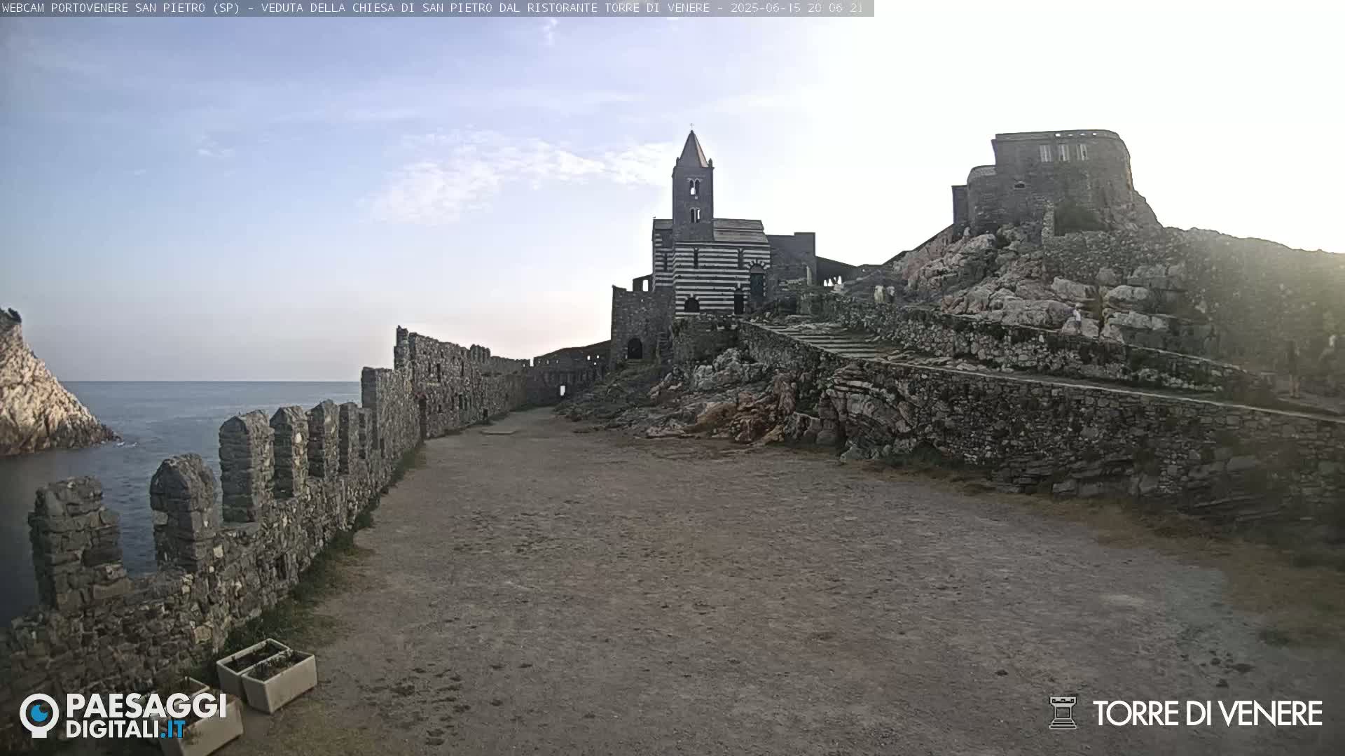 A stone pathway leads to a striped stone church and castle ruins perched atop a rocky cliff overlooking a calm sea under a clear sky.