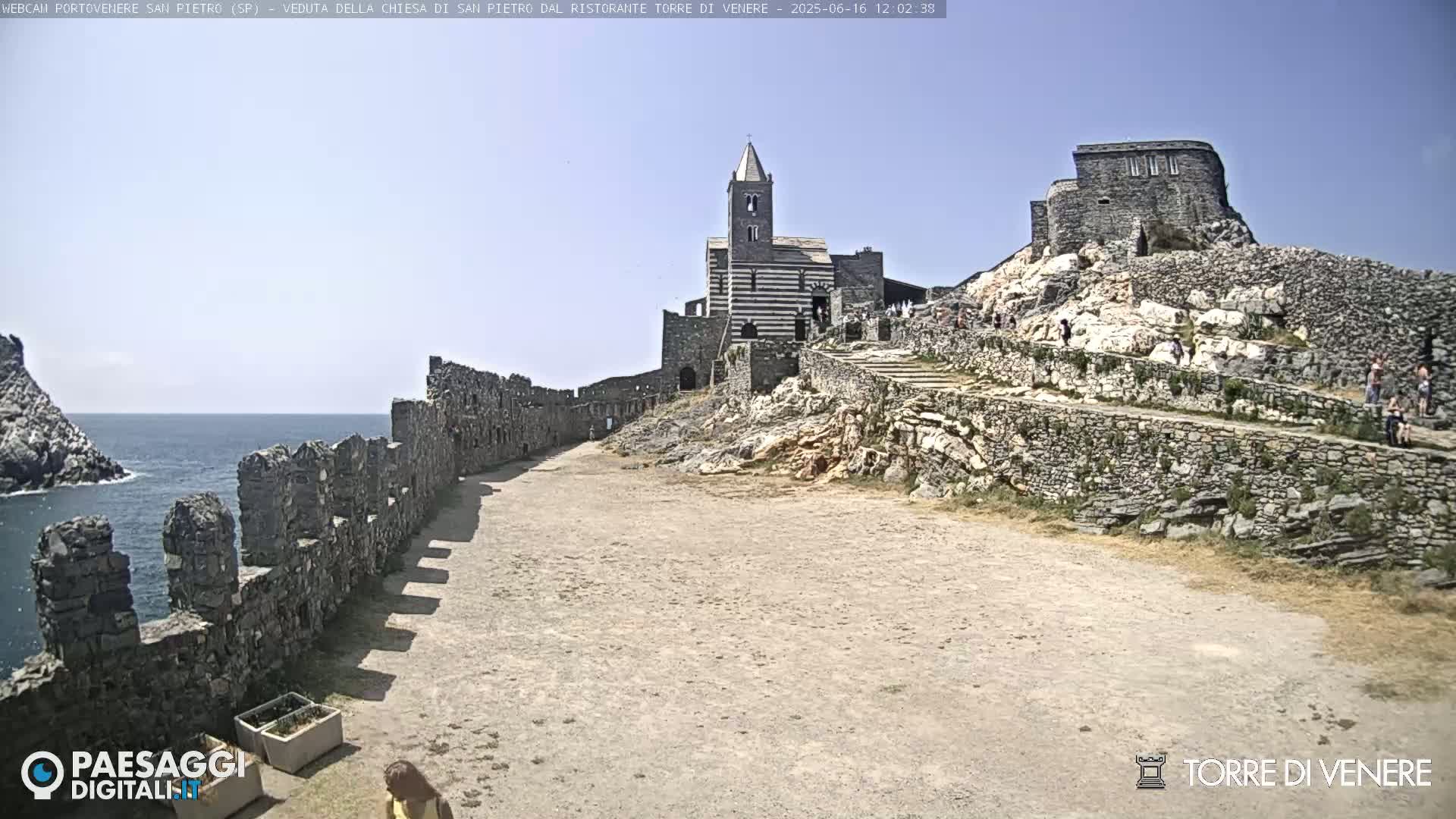 A stone church and castle ruins sit atop a rocky hill overlooking a calm ocean under a clear blue sky.