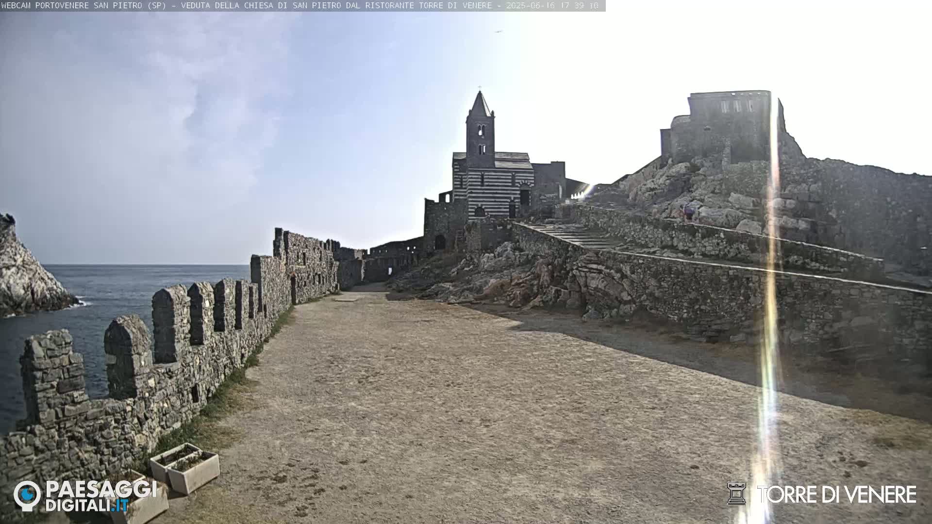 A stone pathway leads to a striped church and castle ruins overlooking a calm ocean under a mostly clear sky.