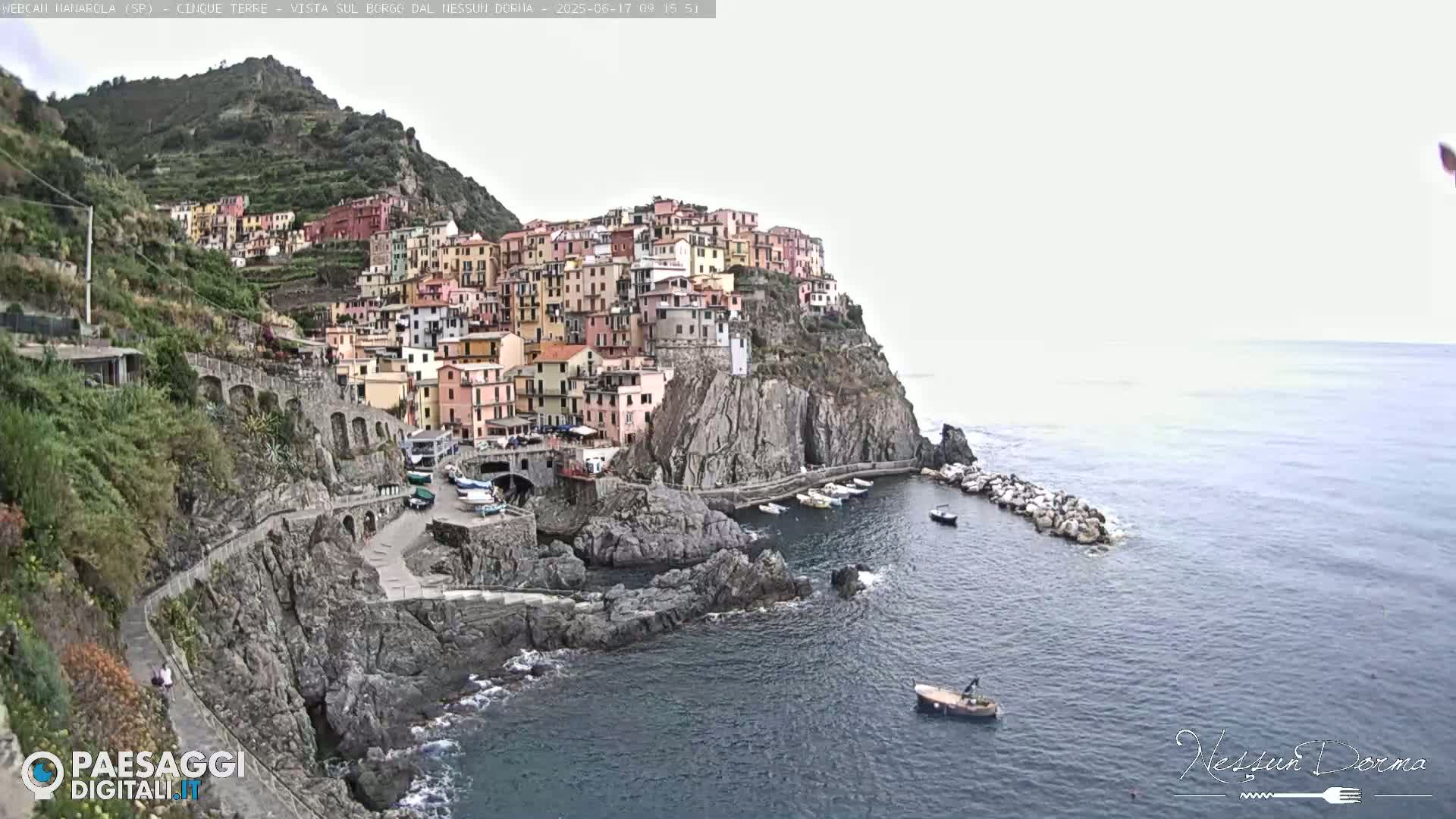 A colorful village clings to a steep, rocky cliffside overlooking a calm sea with a few small boats.