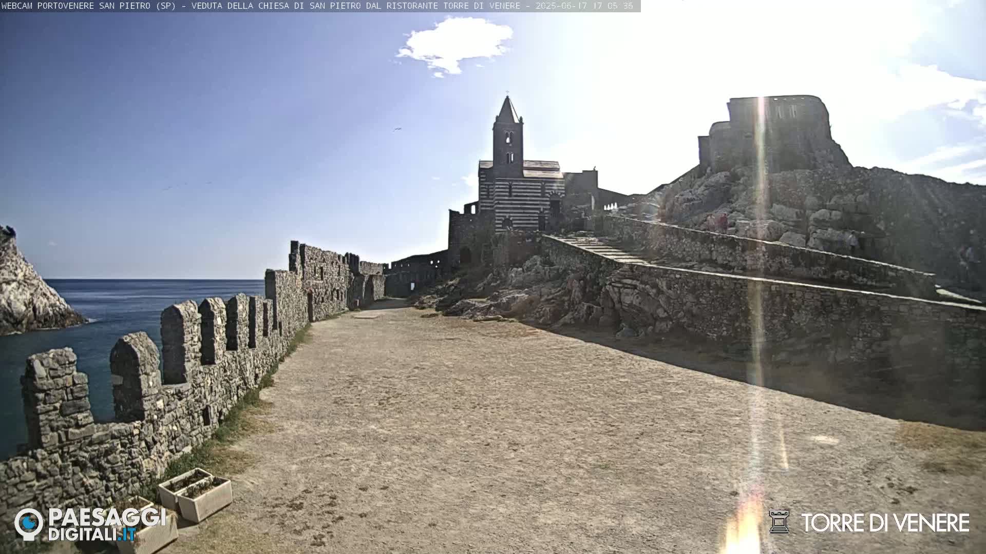 A stone pathway leads to a striped church situated on a rocky outcrop next to a partly ruined stone wall overlooking a calm sea under a sunny sky.