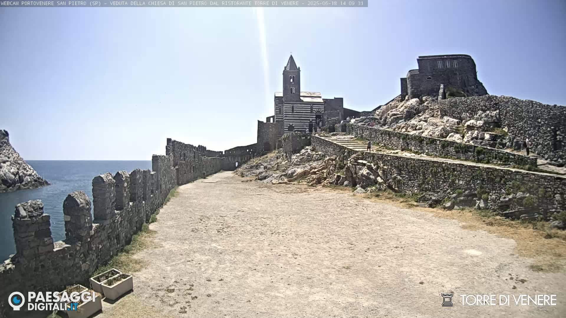 A coastal stone pathway leads to a striped church and castle ruins on a sunny day, overlooking a calm sea.
