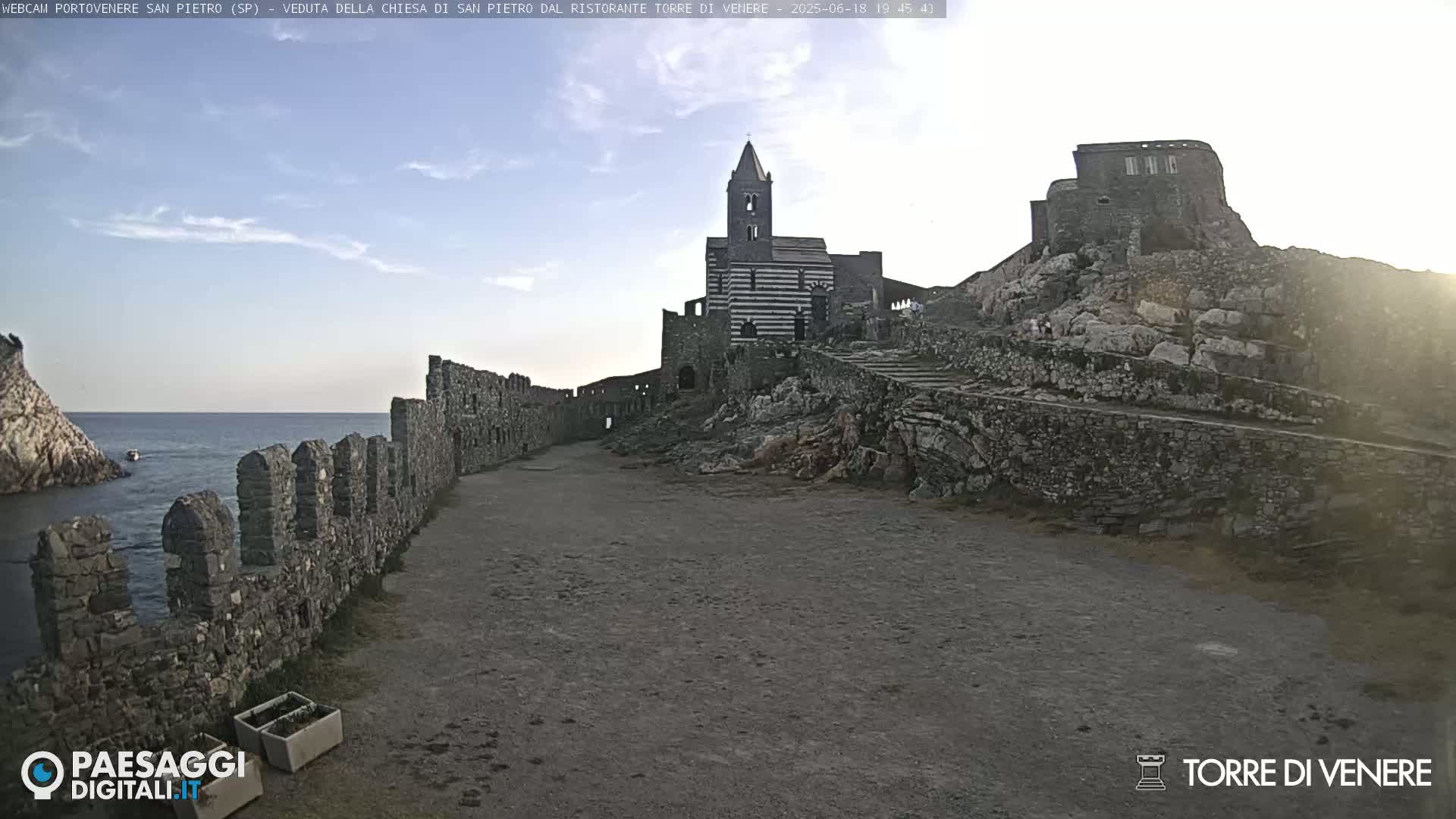 A stone pathway leads toward a striped church and castle ruins perched on a rocky cliff overlooking a calm sea under a mostly clear sky.