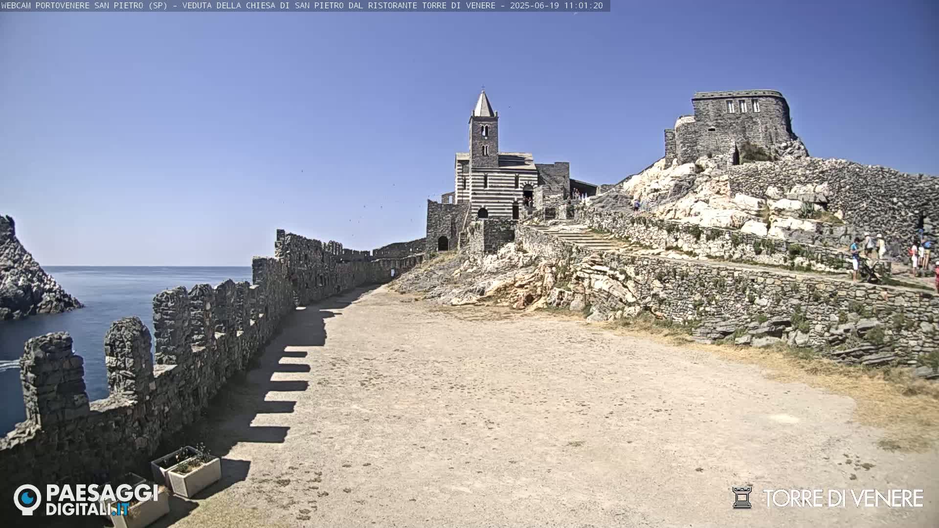 A stone pathway leads to a striped church and stone ruins overlooking a calm ocean under a clear blue sky.