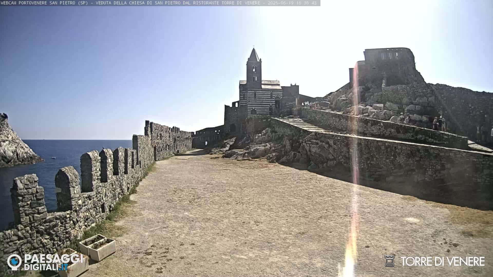 A stone church and castle ruins are situated on a rocky outcrop overlooking a calm sea under a bright, sunny sky.