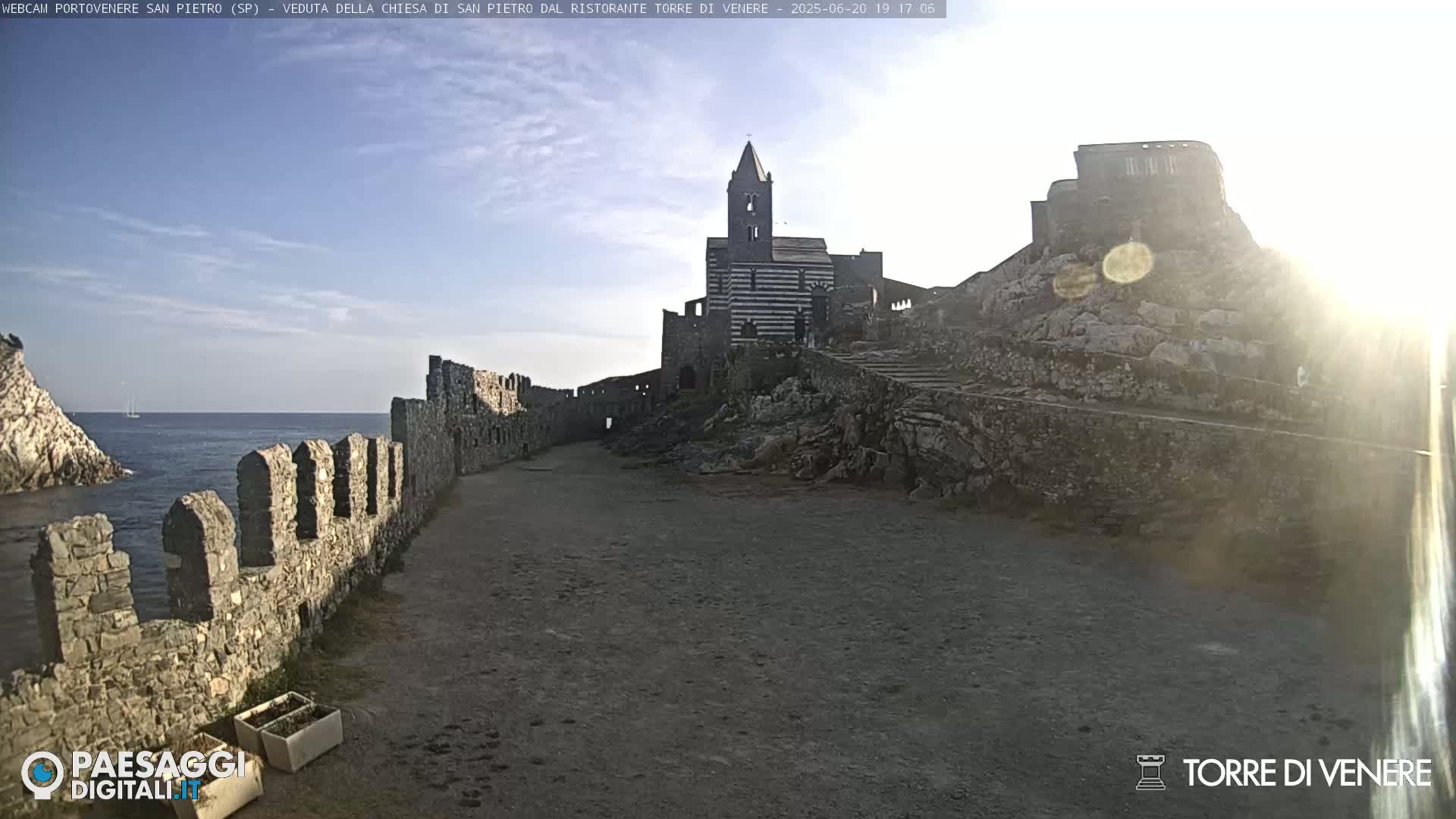 A coastal stone pathway leads to a striped church and castle ruins overlooking a calm sea under a mostly clear sky.