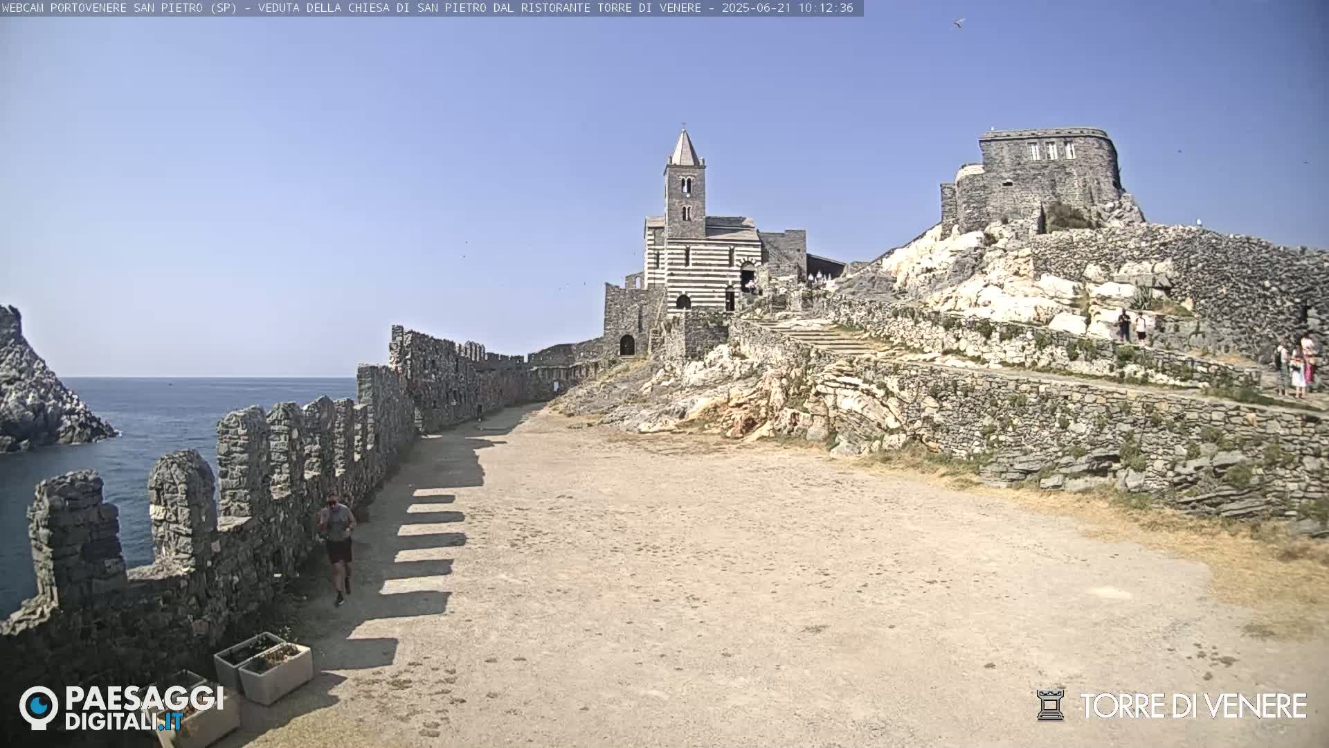 A stone path leads to a striped church and castle ruins perched atop a rocky hill overlooking a calm sea under a clear blue sky.
