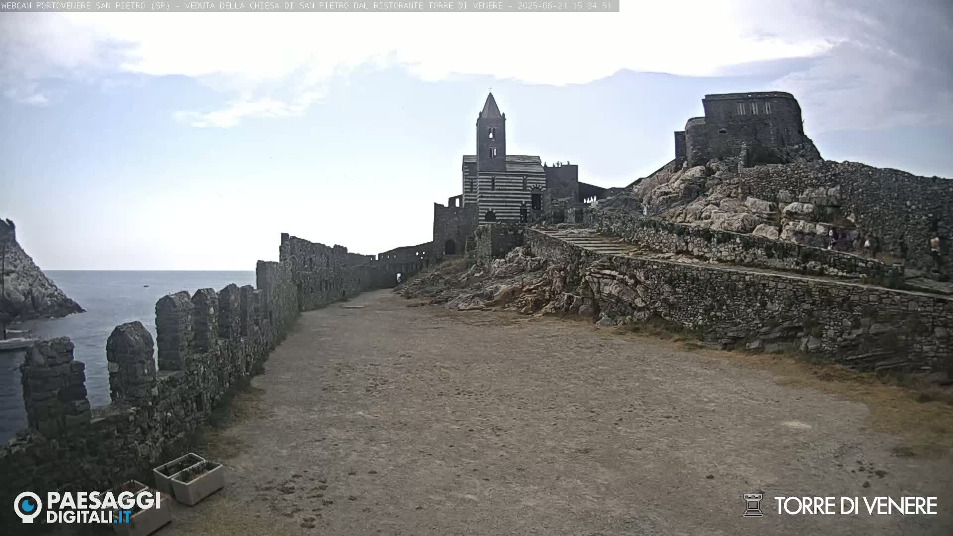 A stone path leads toward a partially ruined church and castle perched on a rocky cliff overlooking a calm sea under a partly cloudy sky.