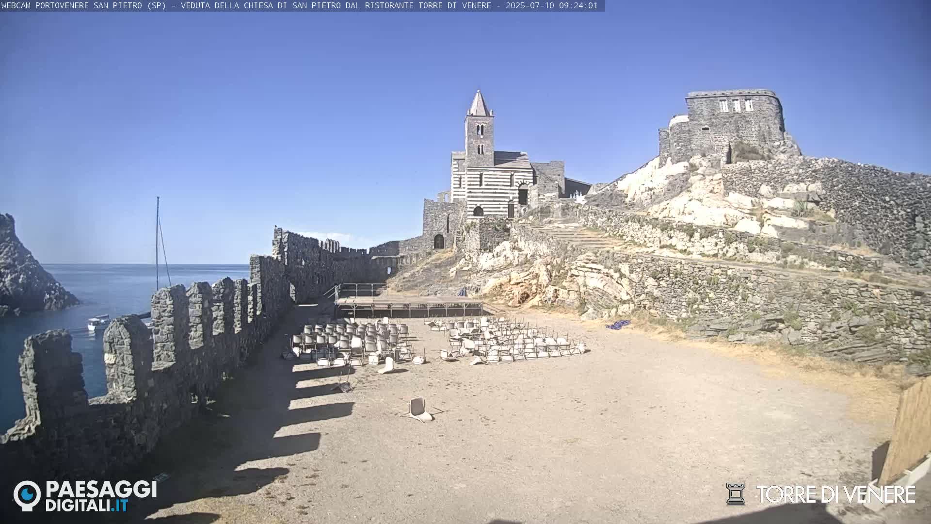 A stone church and castle ruins overlook a sandy area with scattered chairs and a calm ocean under a clear blue sky.