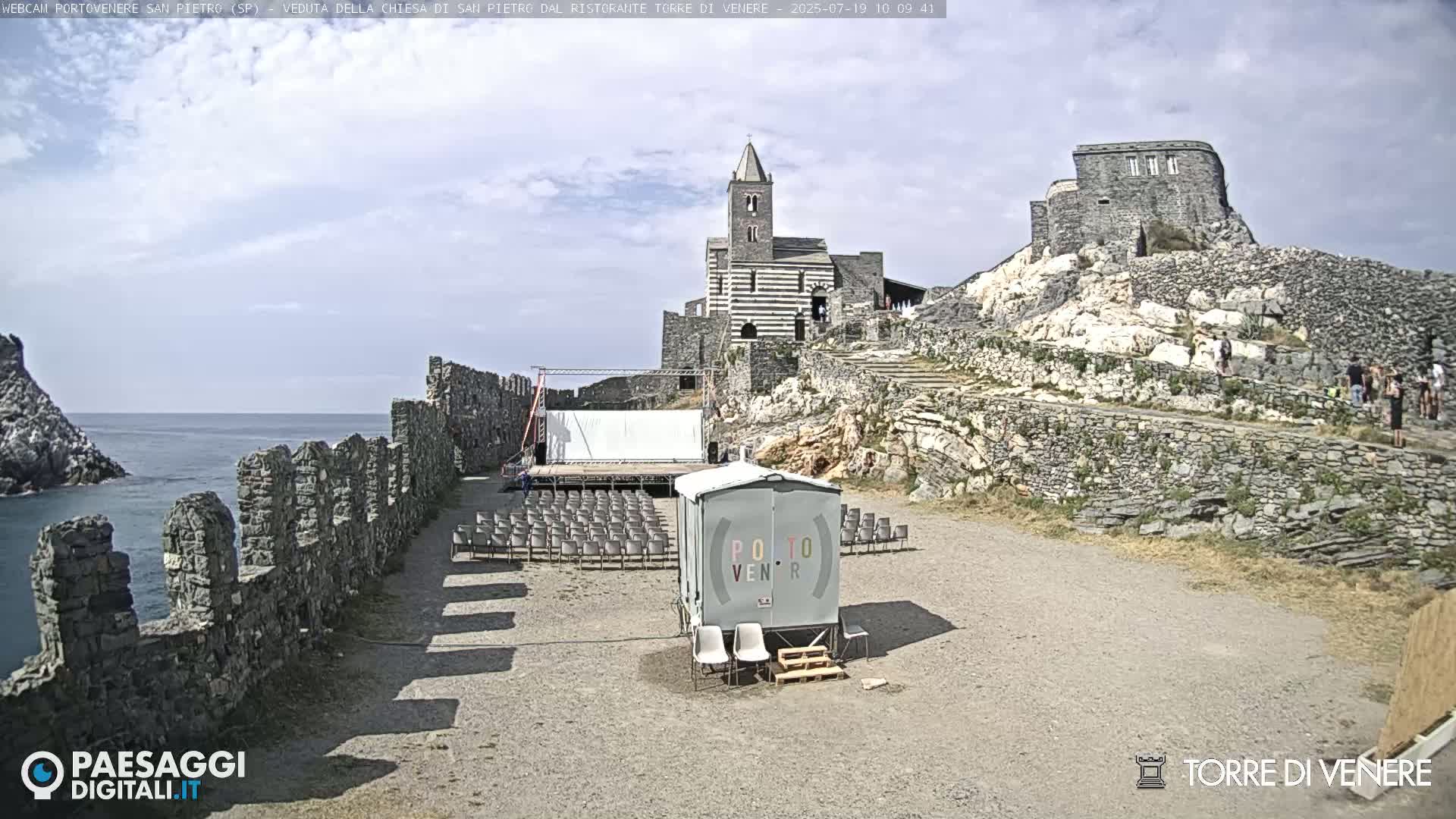 A stone church sits atop a rocky hill overlooking the ocean on a partly cloudy day, with a temporary stage and seating area set up in the foreground.