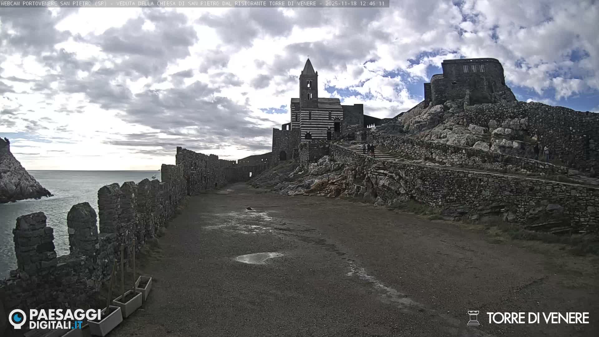 A historic church with a striped facade, illuminated by blue lights, stands on a rocky hill at night under clear skies, with ancient stone structures surrounding it and a path lit by yellow artificial lights in the foreground.