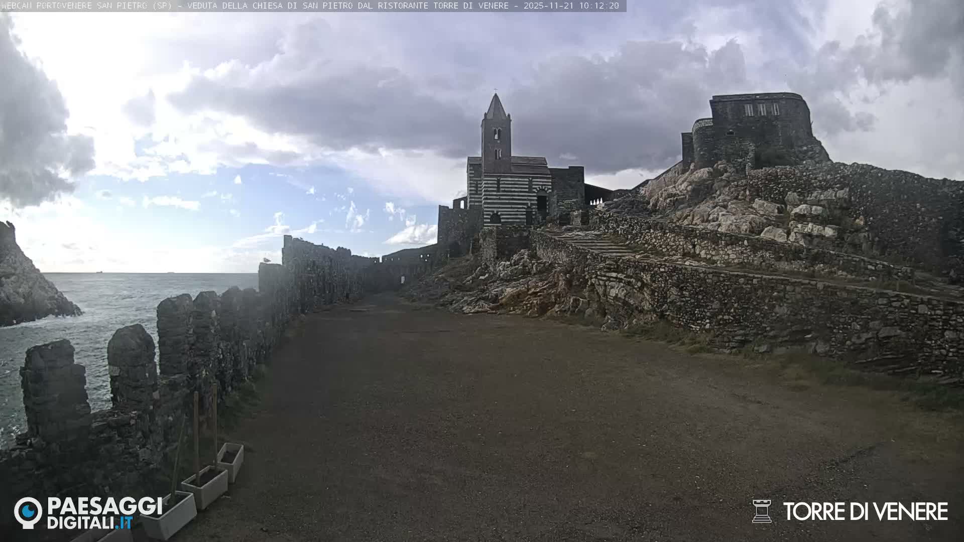 Historic stone fortifications line a rocky coast next to a choppy sea, leading to a distinctive striped church with a bell tower and other ancient buildings perched on cliffs under a partly cloudy sky.