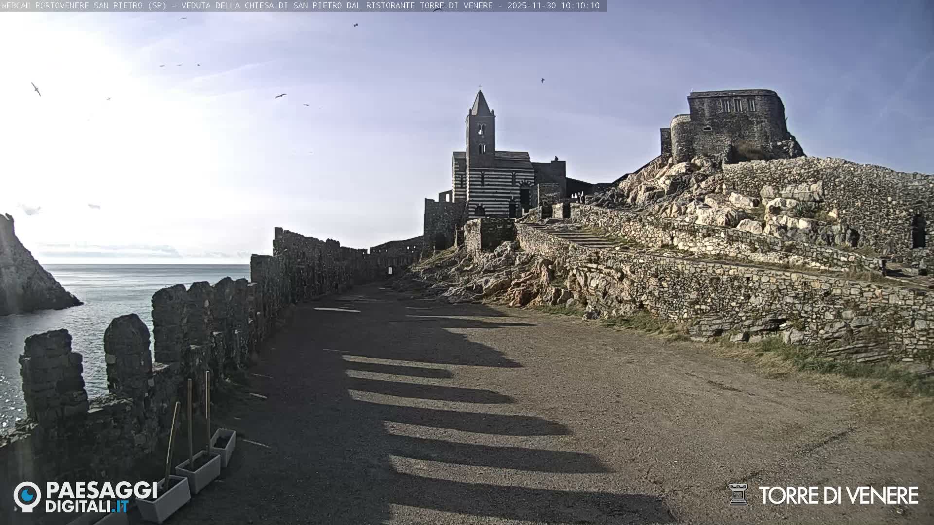 Under a bright, partly cloudy sky, a wide path leads along a rugged coastline featuring ancient stone walls and structures, including a distinctive striped church, overlooking a calm sea.