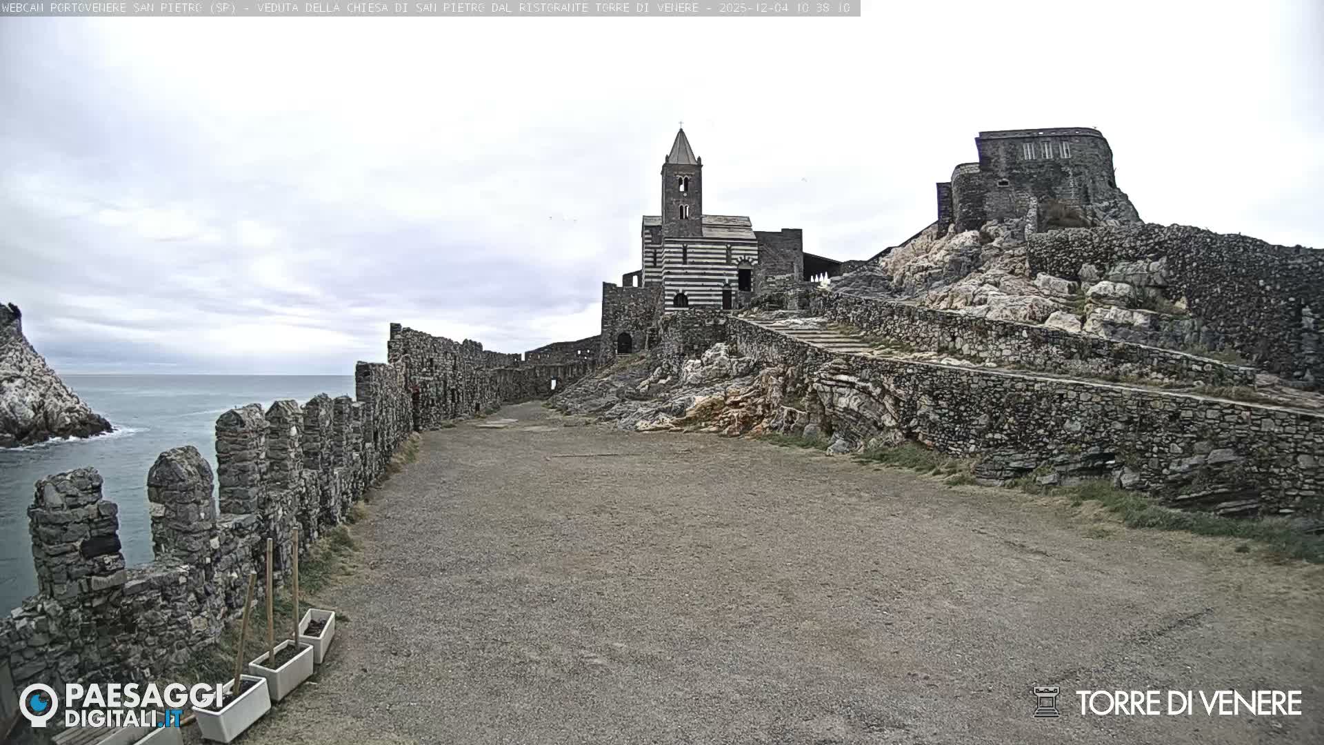 A historic stone church with a striped facade and bell tower stands on a rocky promontory, surrounded by ancient stone walls and a more ruinous fortress on a hill, overlooking a calm sea under a cloudy, overcast sky.