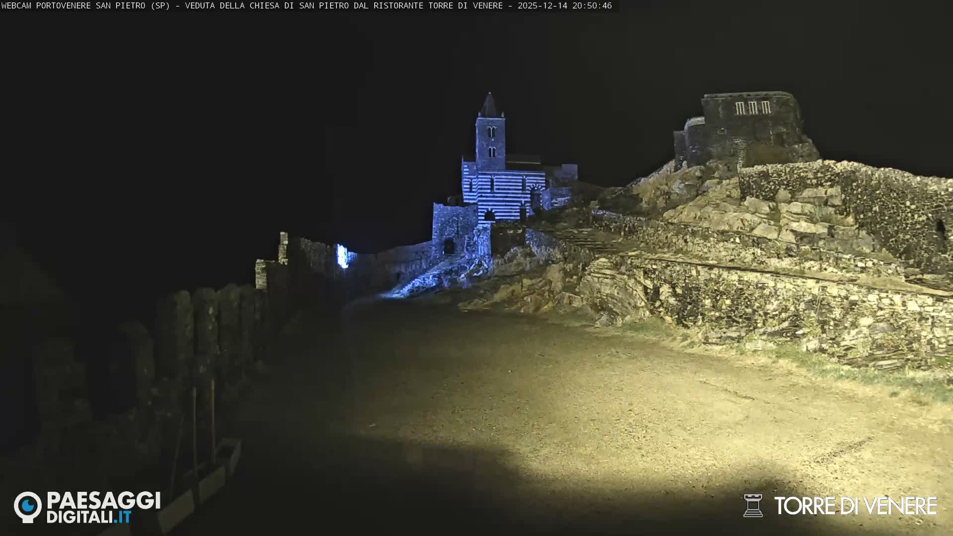 A historic stone church with a striped facade and bell tower stands on a rocky promontory, surrounded by ancient stone walls and a more ruinous fortress on a hill, overlooking a calm sea under a cloudy, overcast sky.