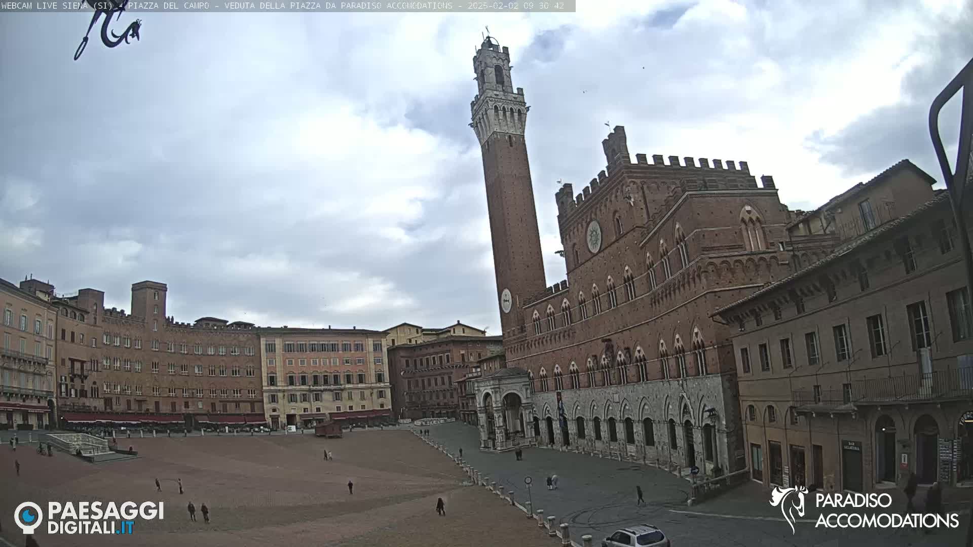A large, brick piazza in Siena, Italy, with a tall tower and several historical buildings, under a mostly cloudy sky.