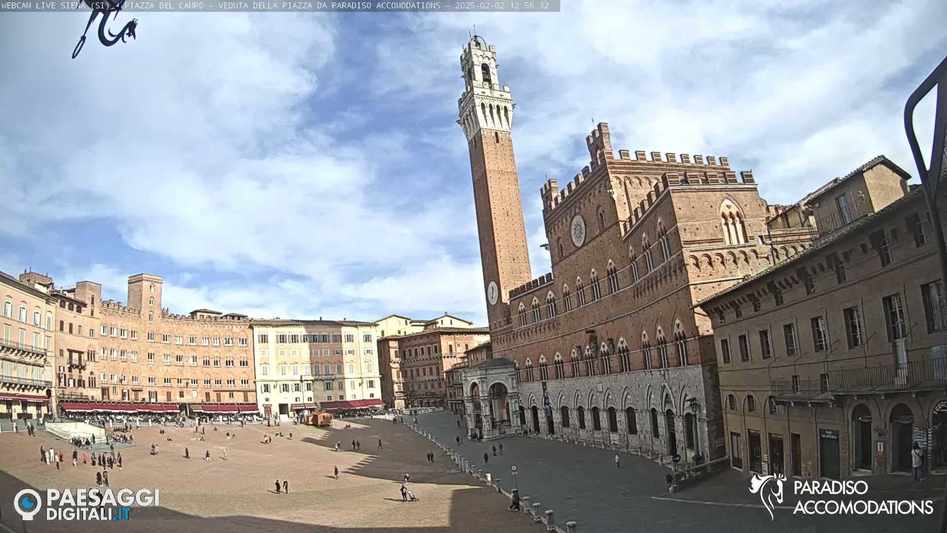A large, brick public square in a European city, under a partly cloudy sky, features a tall, slender tower and a large palace-like building.