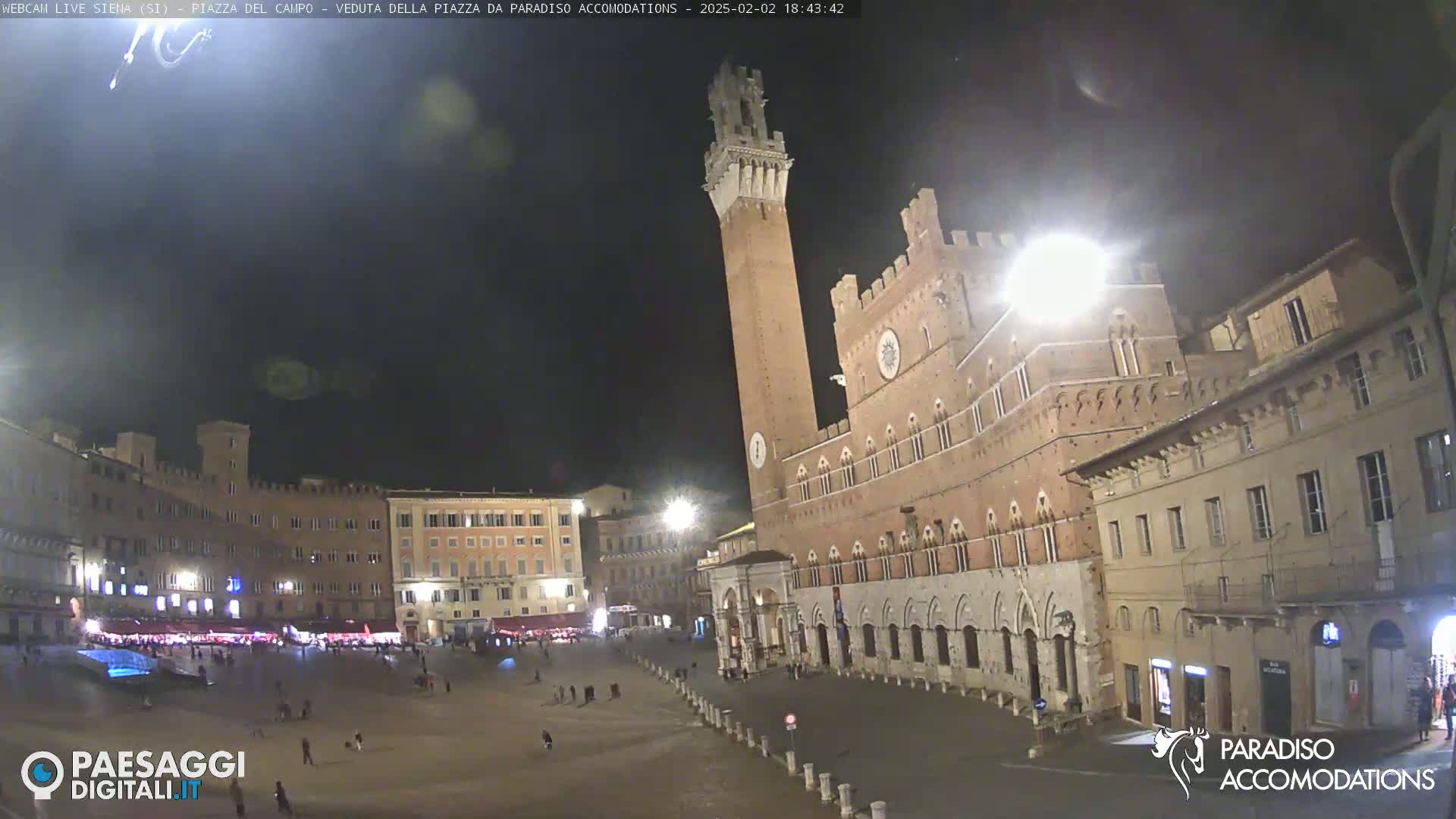 A large, mostly empty plaza at night, surrounded by historic brick buildings, with a tall tower prominently featured in the center.