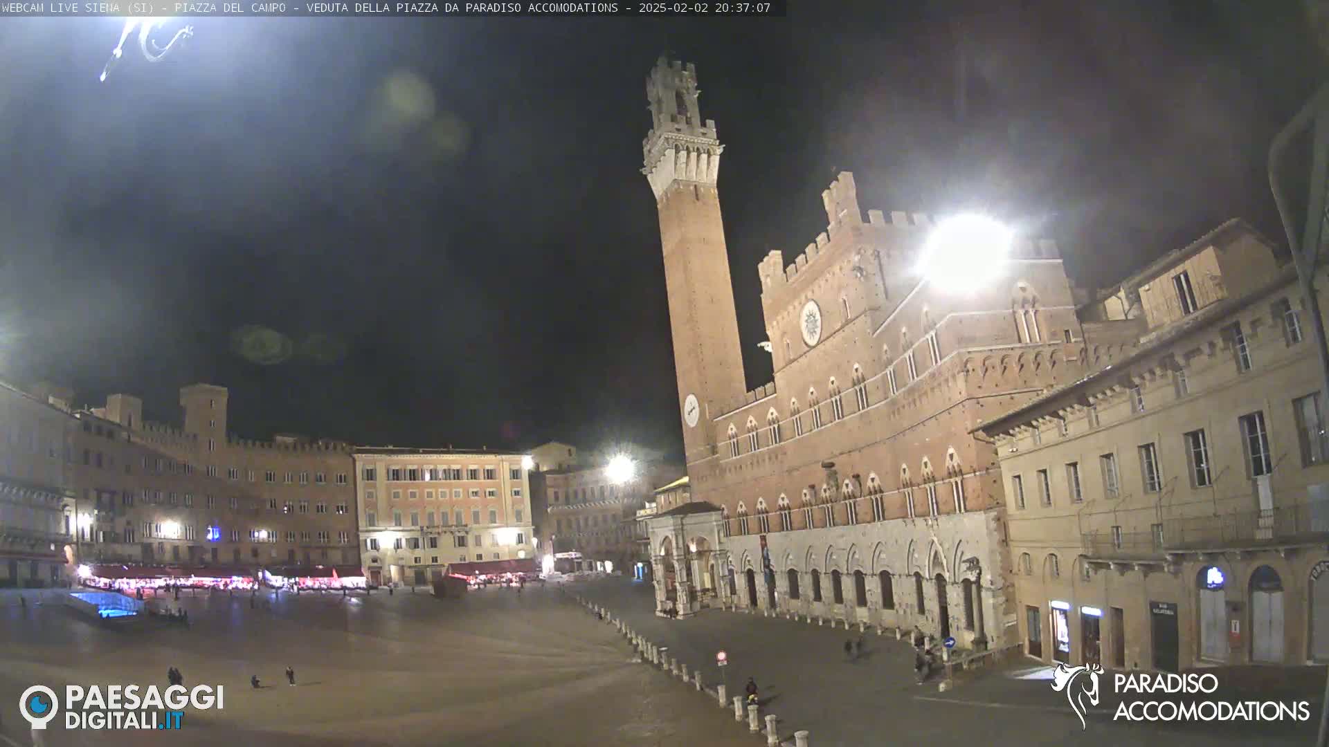 A nighttime view of a large, mostly empty plaza surrounded by historic brick buildings, one with a tall tower, under a dark sky.