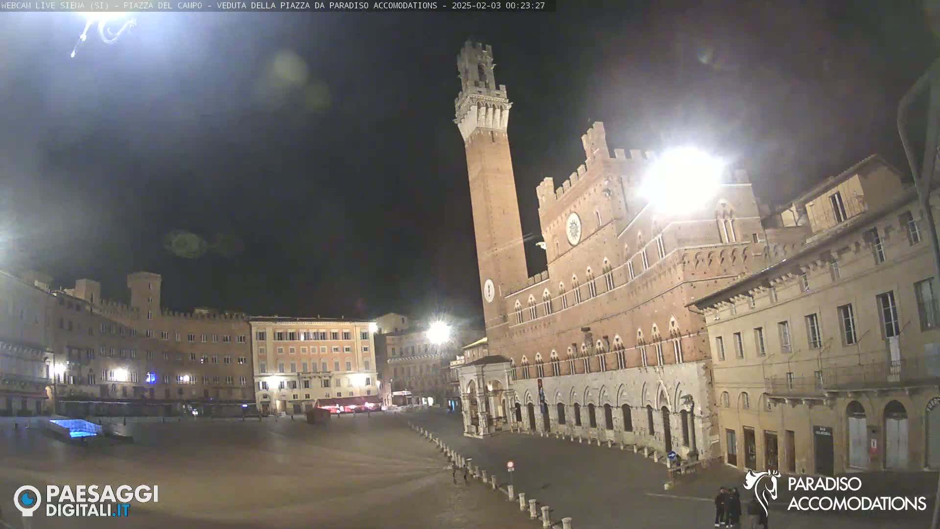 A nighttime view of a large, empty plaza with historical buildings, including a tall brick tower, under a dark, mostly clear sky.