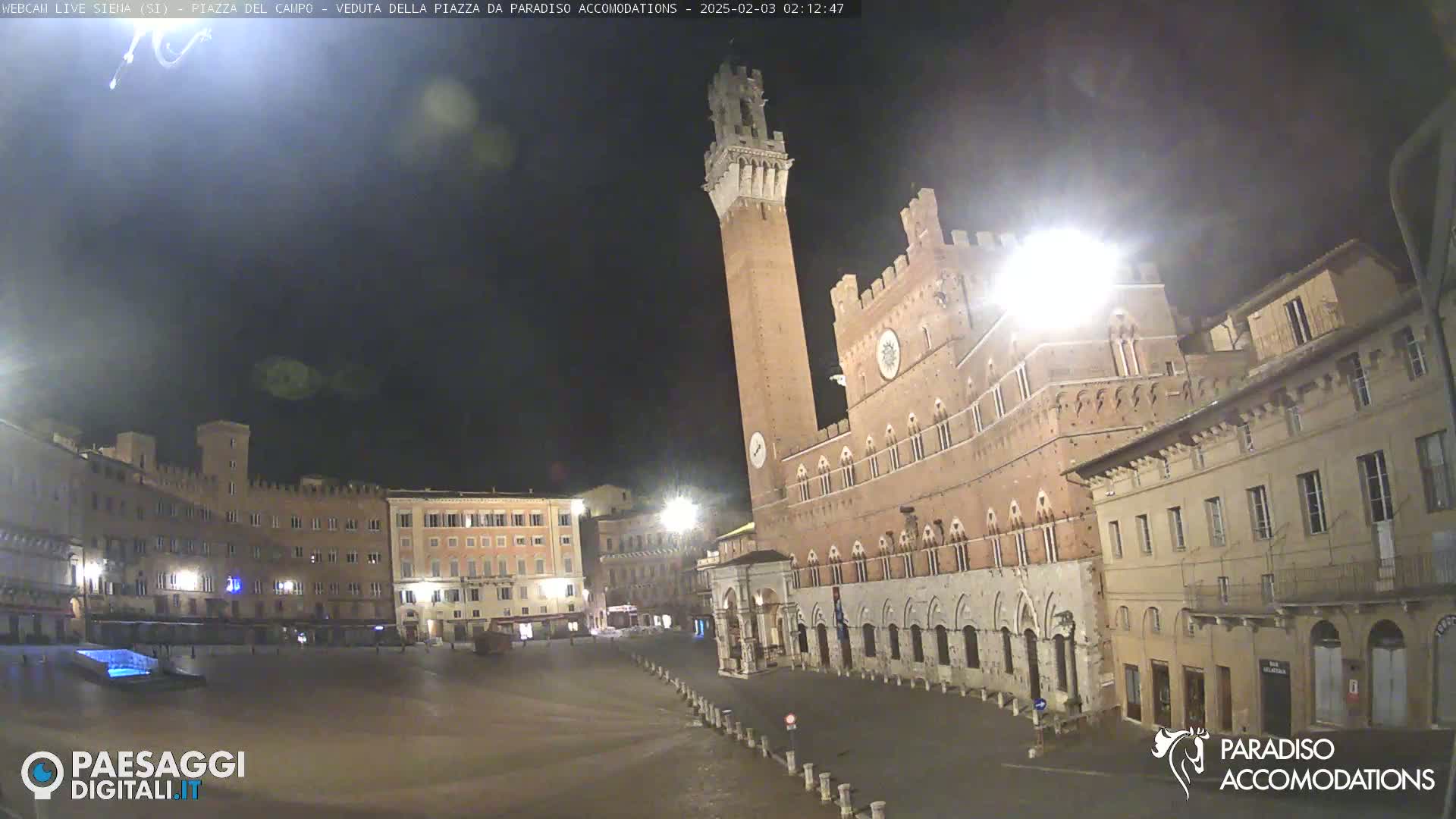 A mostly empty, brightly lit town square at night, dominated by a tall, brick tower and adjacent building, under a dark, clear sky.