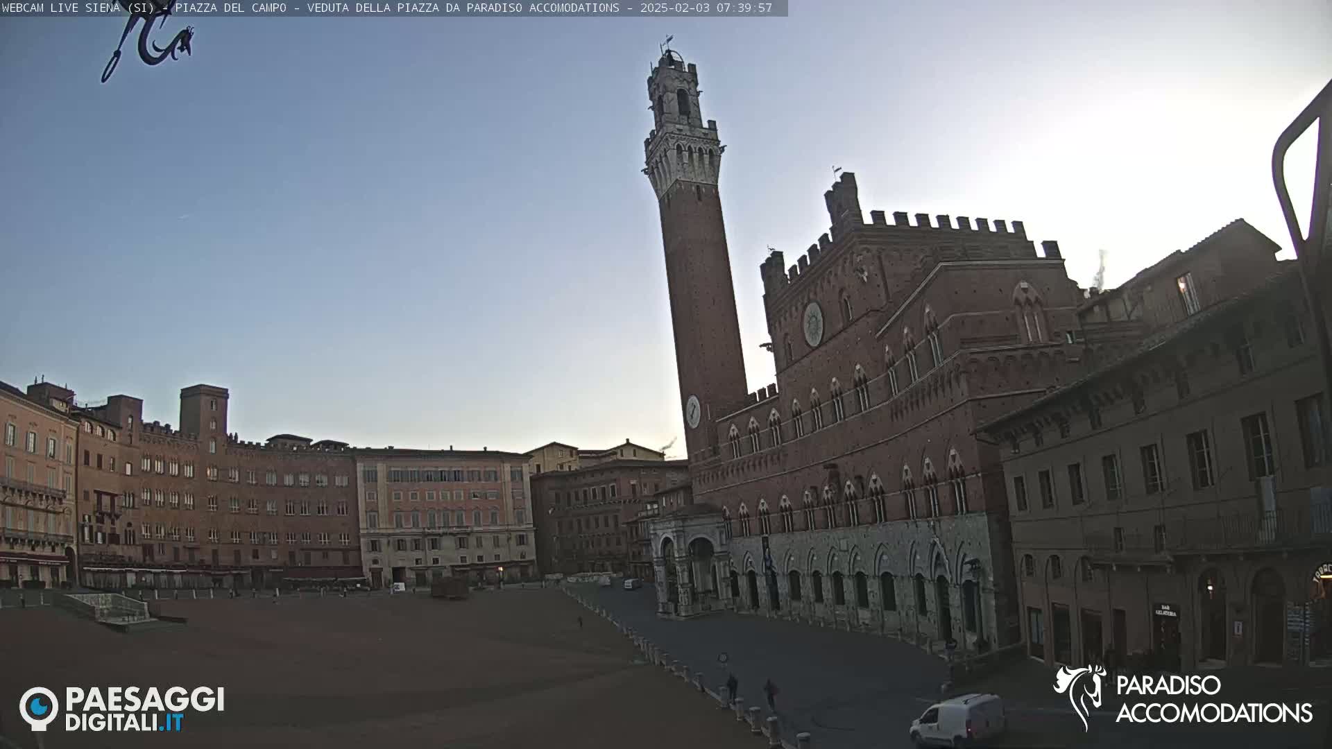 A large, brick piazza in a town is visible at dawn under a clear sky; a tall tower stands prominently near a large, ornate building.