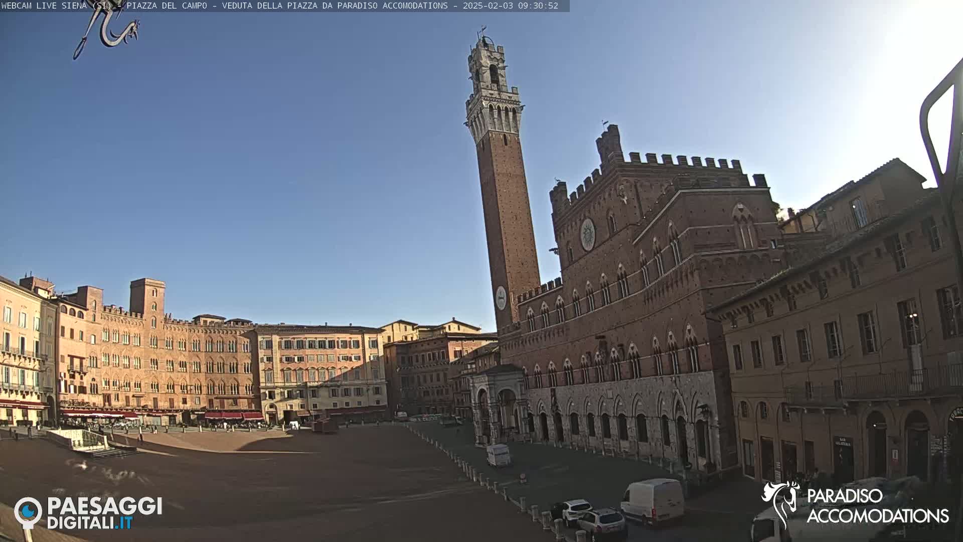 A large, brick public square is shown on a sunny day, surrounded by historic buildings, including a tall tower and a palace.