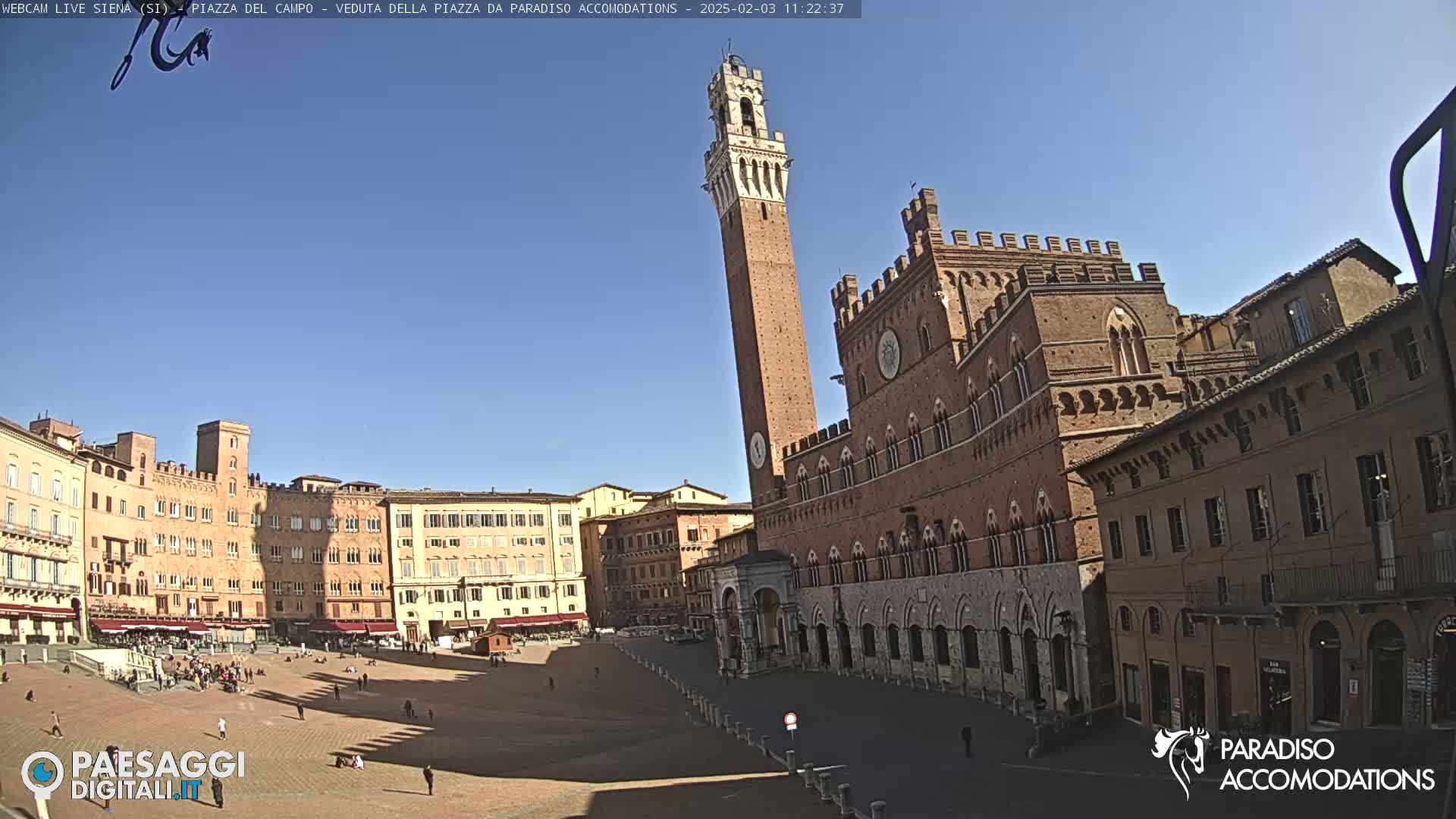 A large, brick piazza in bright sunlight is surrounded by medieval-style buildings, including a tall tower.