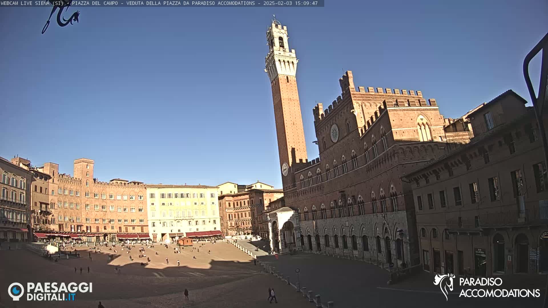 A large, brick public square in Italy is mostly empty under a clear blue sky, with a tall tower and several large buildings surrounding it.