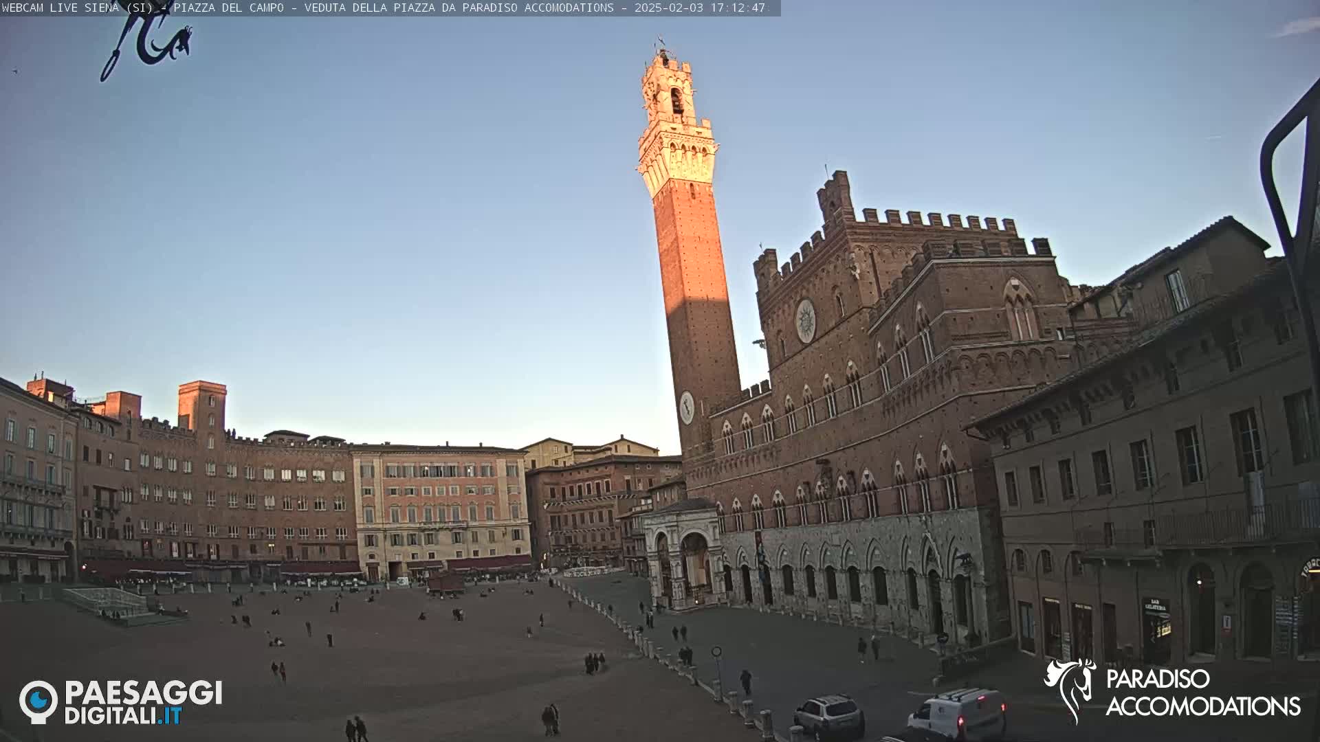 A large, brick piazza in Siena, Italy, is shown at sunset under a clear sky.