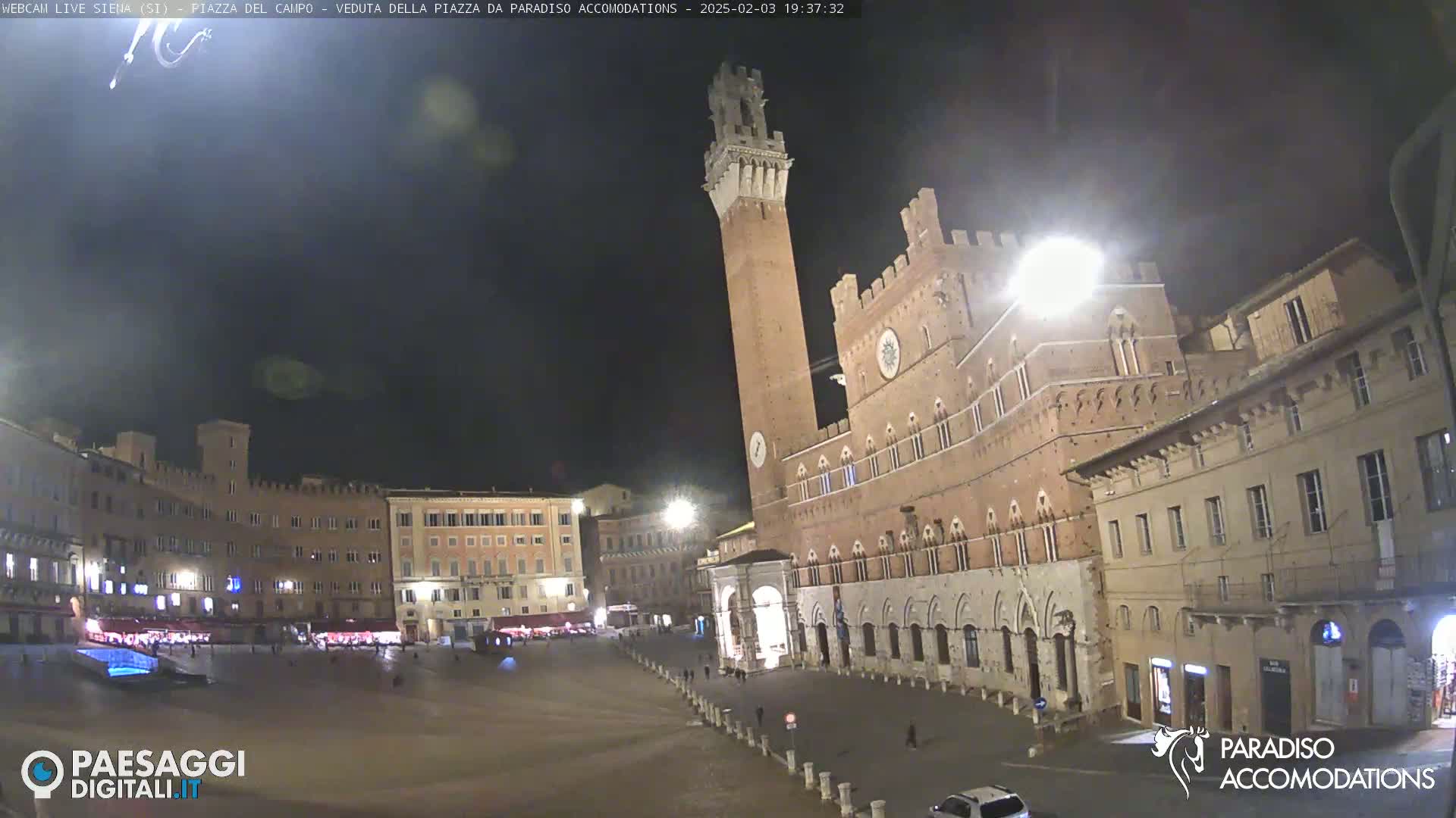 A nighttime view of a large, empty town square in Italy, surrounded by historic brick buildings, one prominently featuring a tall tower.