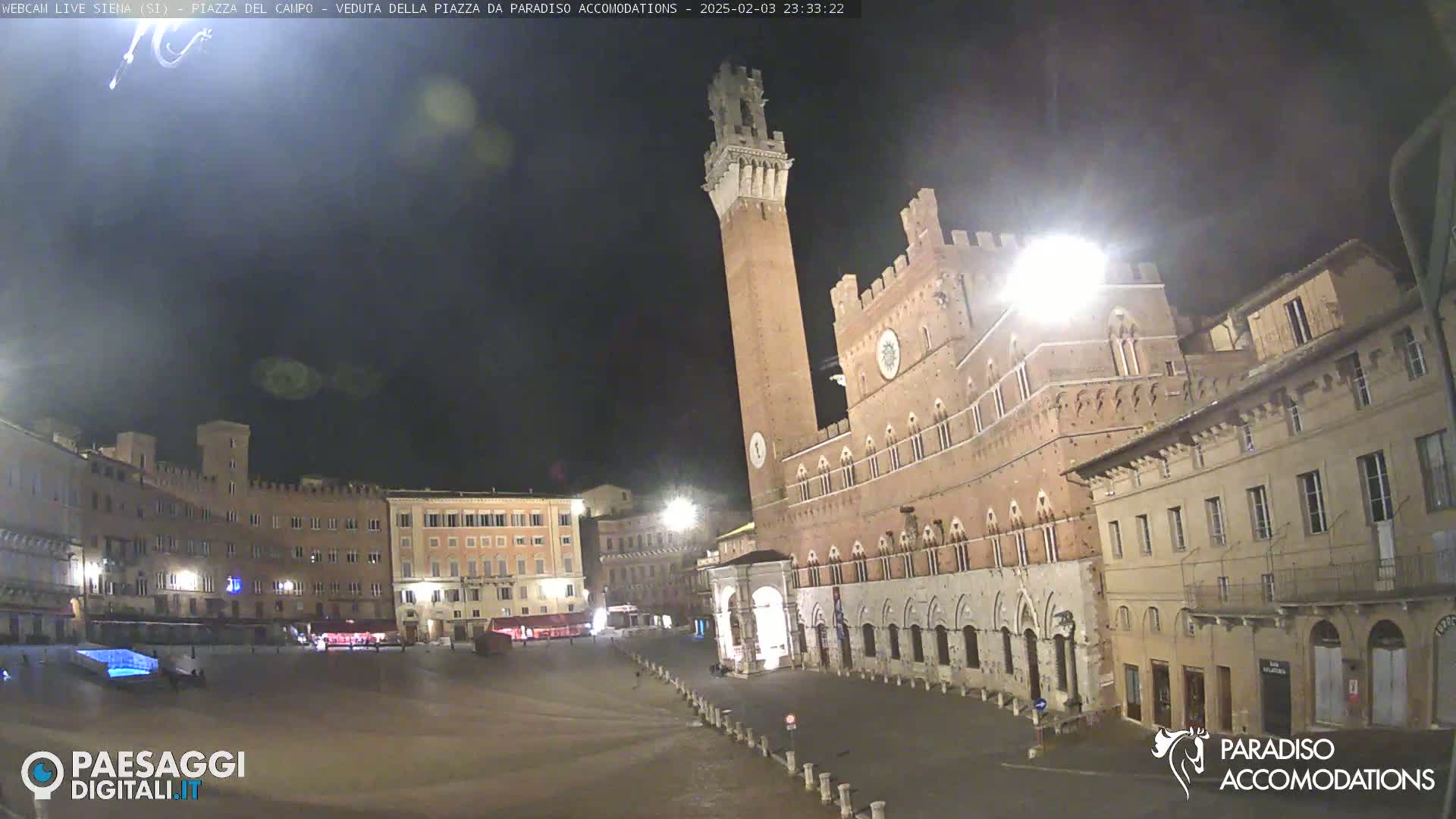 A large, mostly empty, square at night is surrounded by historic brick buildings, one with a tall tower, under a dark, clear sky.