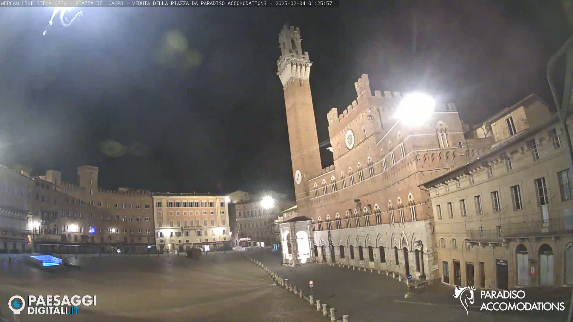 A large, empty plaza at night, featuring a tall brick tower and surrounding historic buildings under a dark sky.