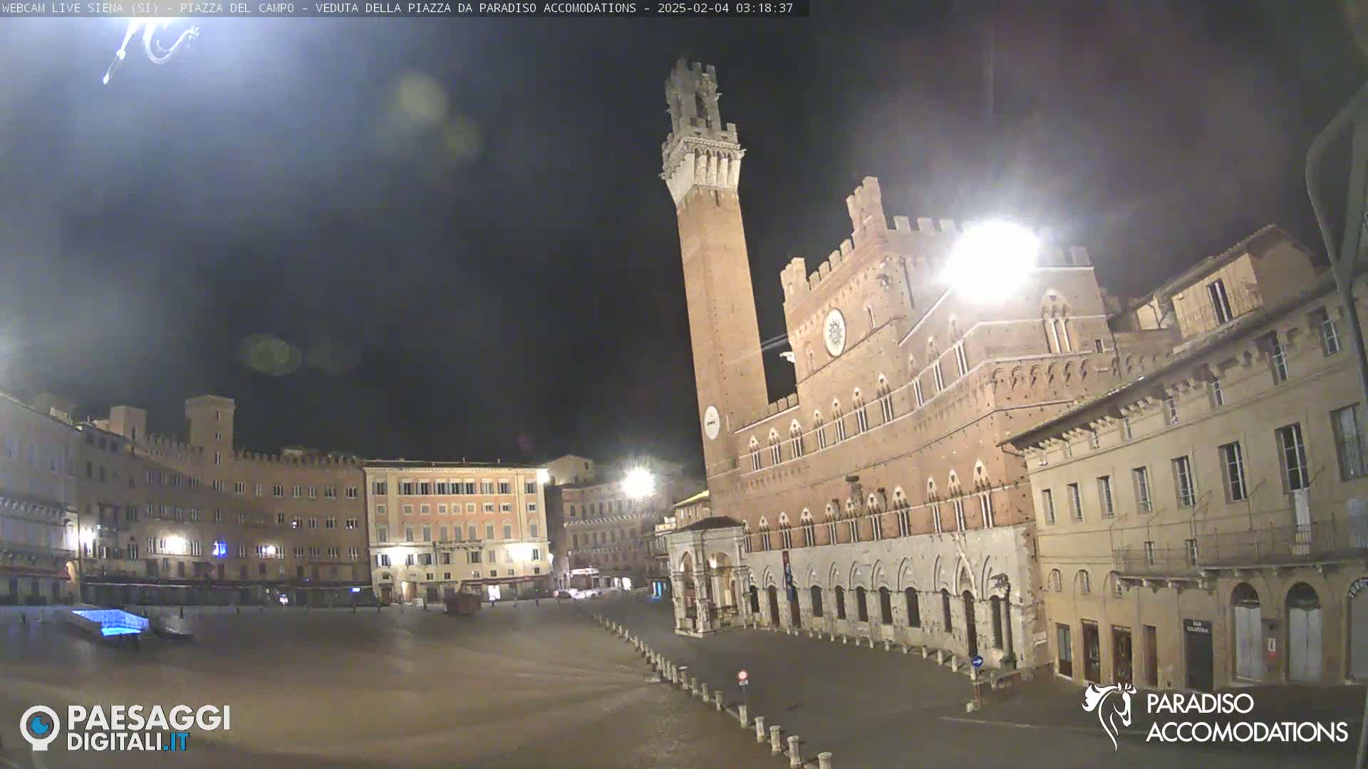 A nighttime view of a large, empty plaza with historical buildings, including a tall tower, under a dark, clear sky.